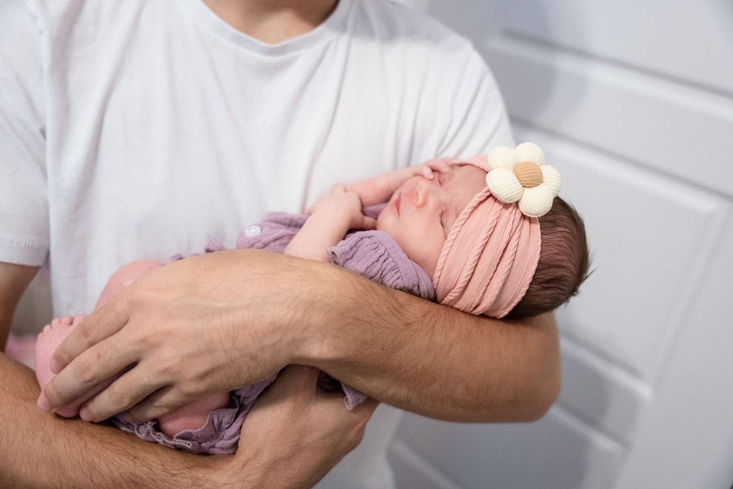 Father in white t-shirt cradling sleeping newborn baby girl wearing pink swaddle and floral headband.