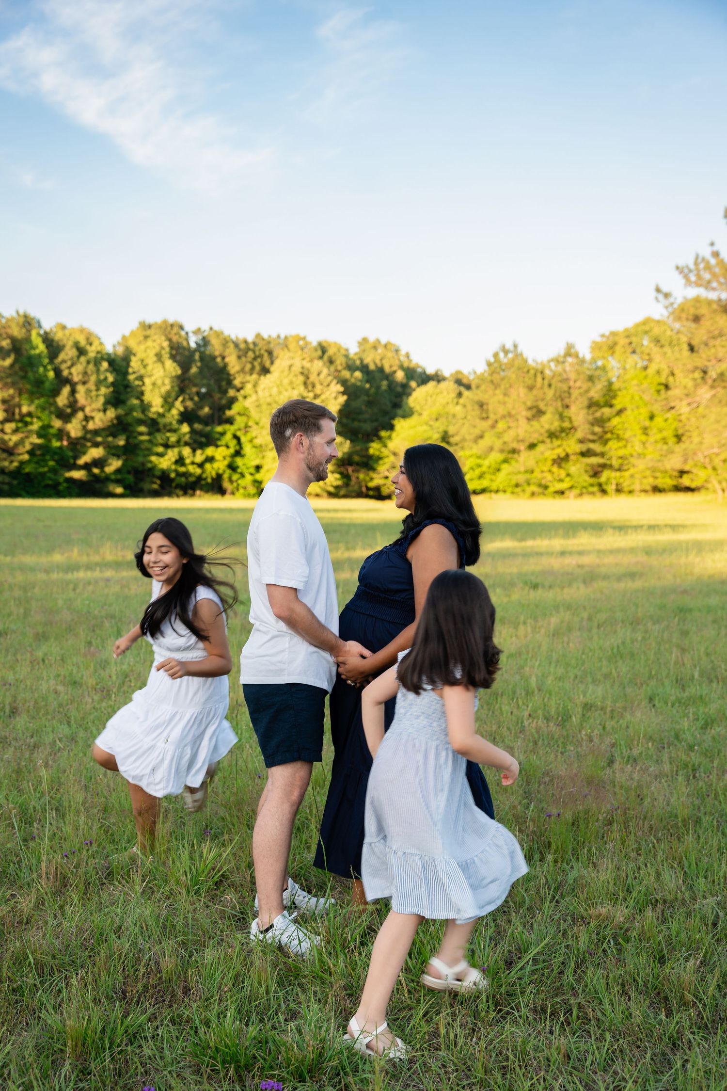 Happy family in a green field, pregnant mother and father with two daughters in white dresses during golden hour.
