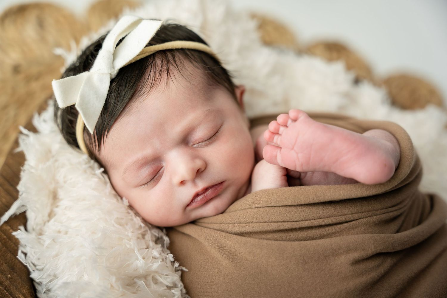 A sleeping newborn baby swaddled in brown fabric lies on a white textured blanket during a professional photo session.