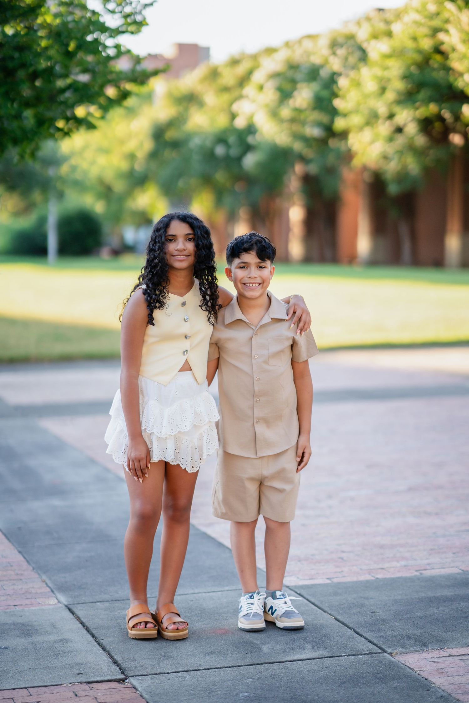 A smiling teenage girl and young boy pose together on a sunny outdoor pathway.