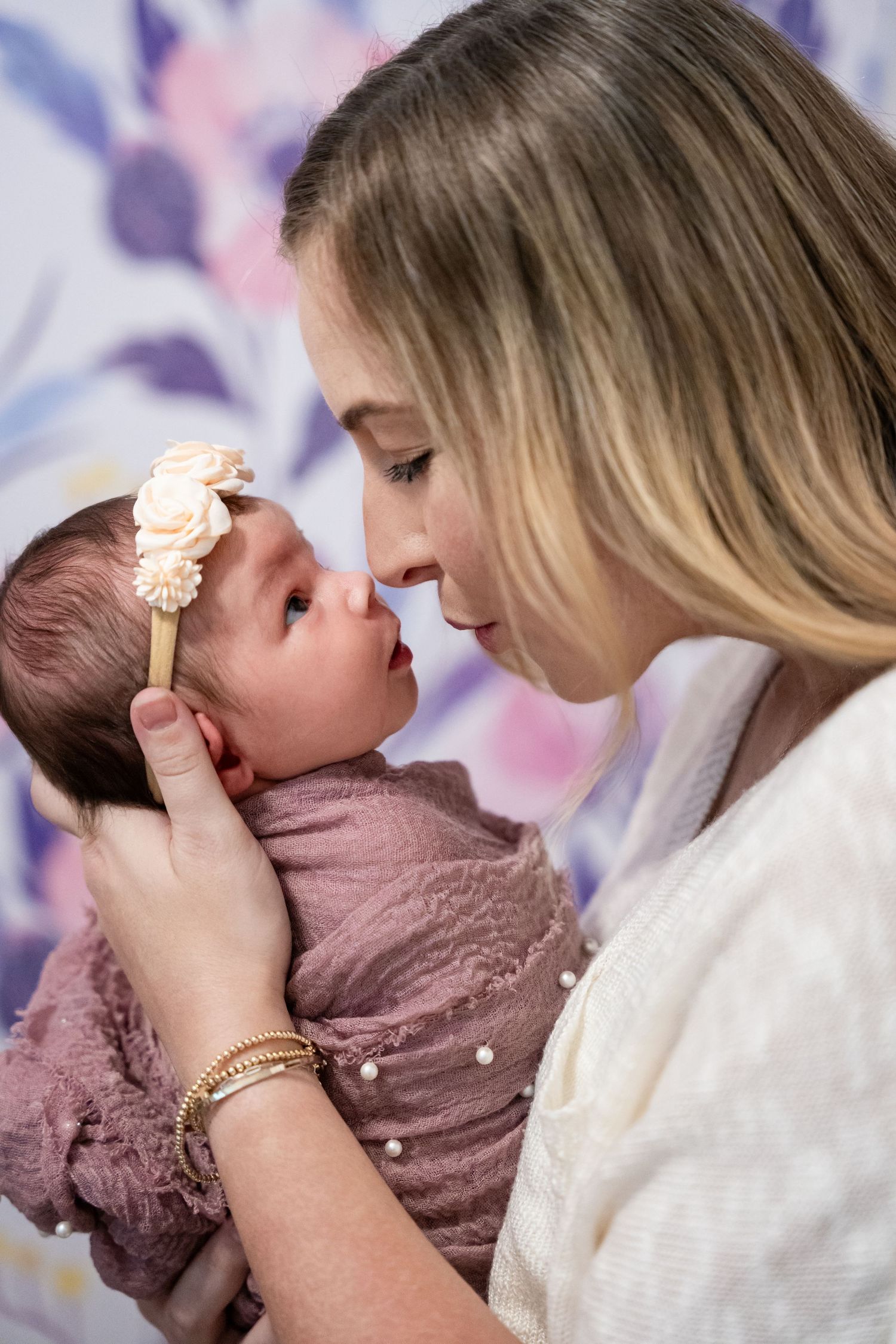 Young mother tenderly holds newborn baby girl wearing floral headband, wrapped in pink swaddle, sharing a sweet nose-to-nose moment.