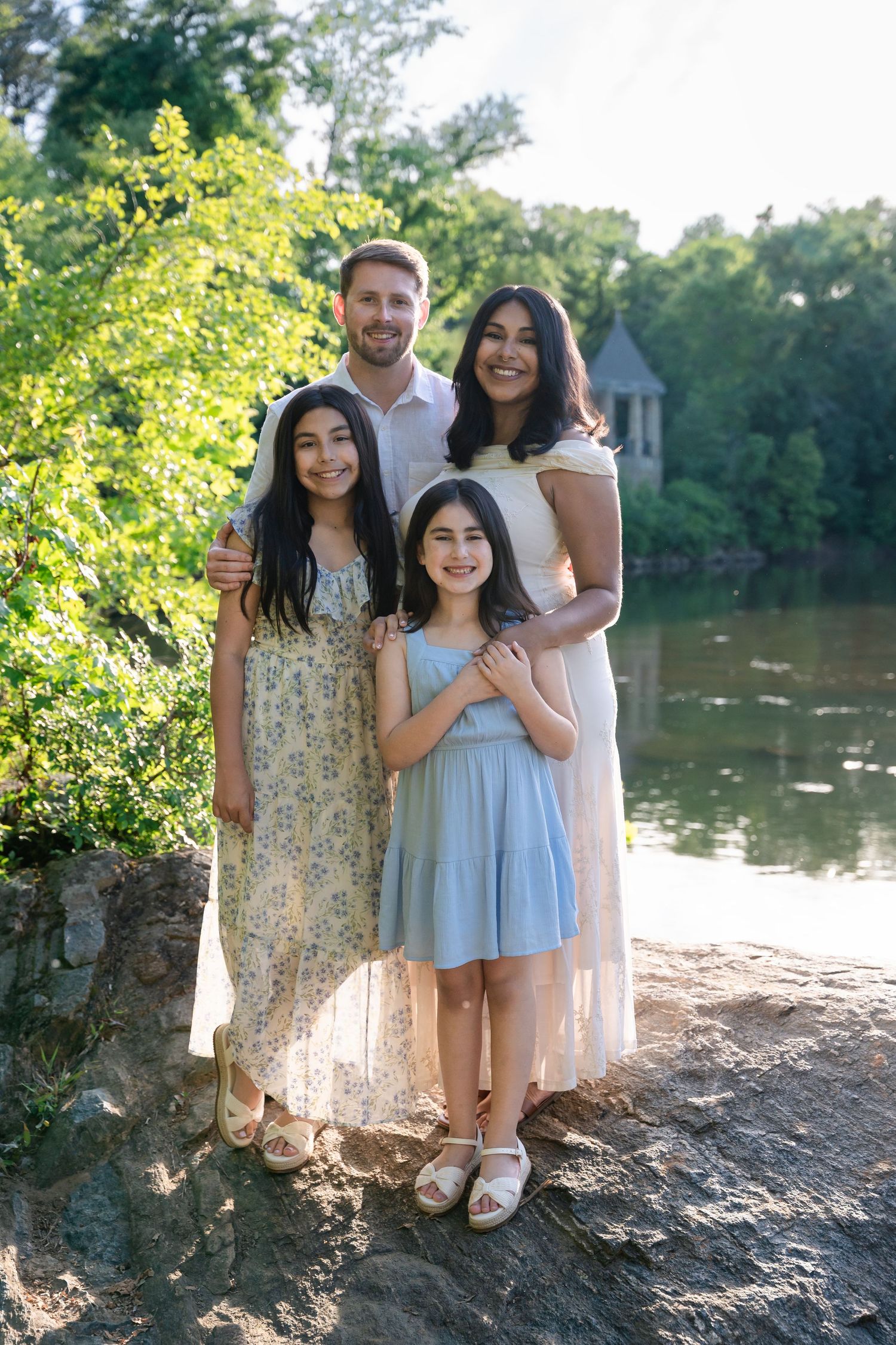 Happy family of four posing by a serene pond surrounded by lush greenery on a sunny day.