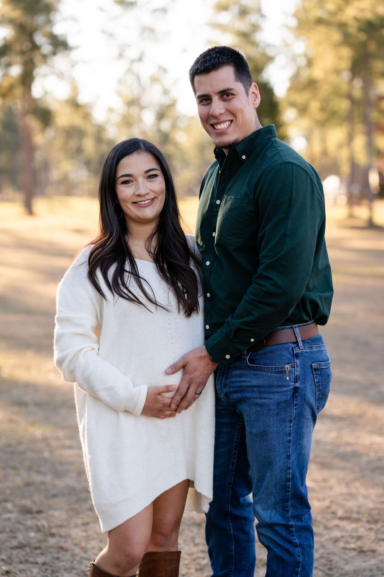 A couple poses together in casual attire against a backdrop of pine trees in warm afternoon light.
