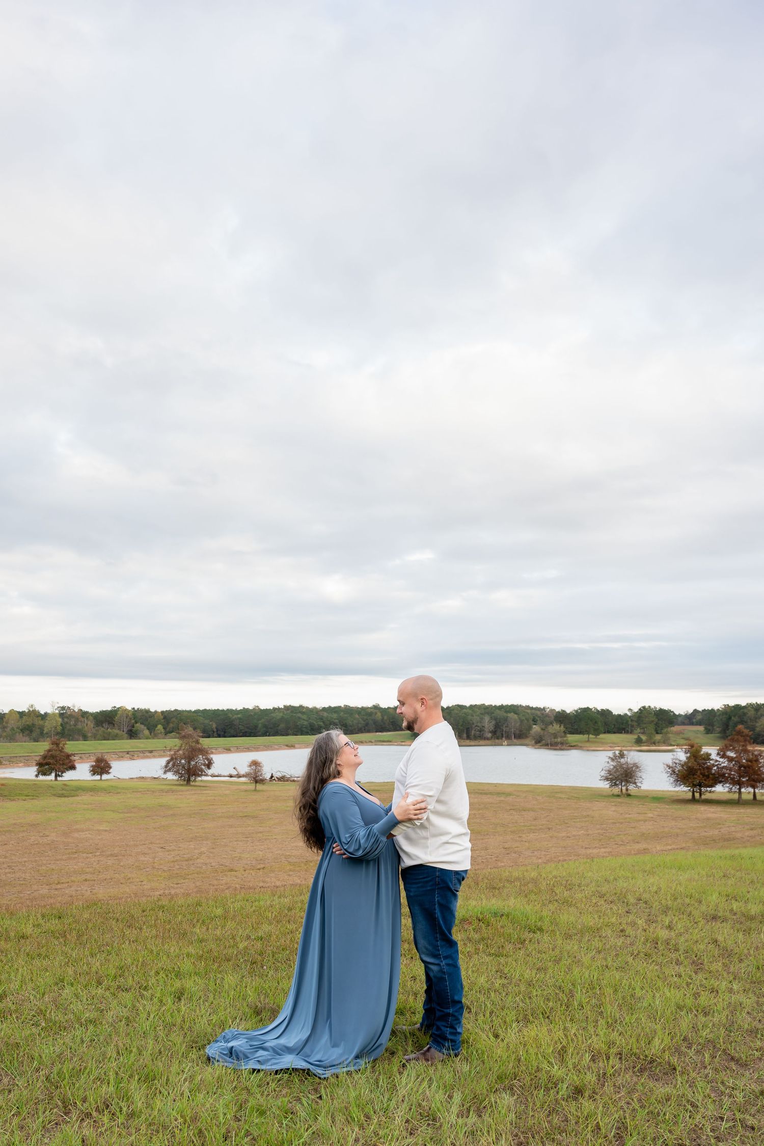 Couple in casual clothing posing together on grassy field with lake and cloudy sky in background.