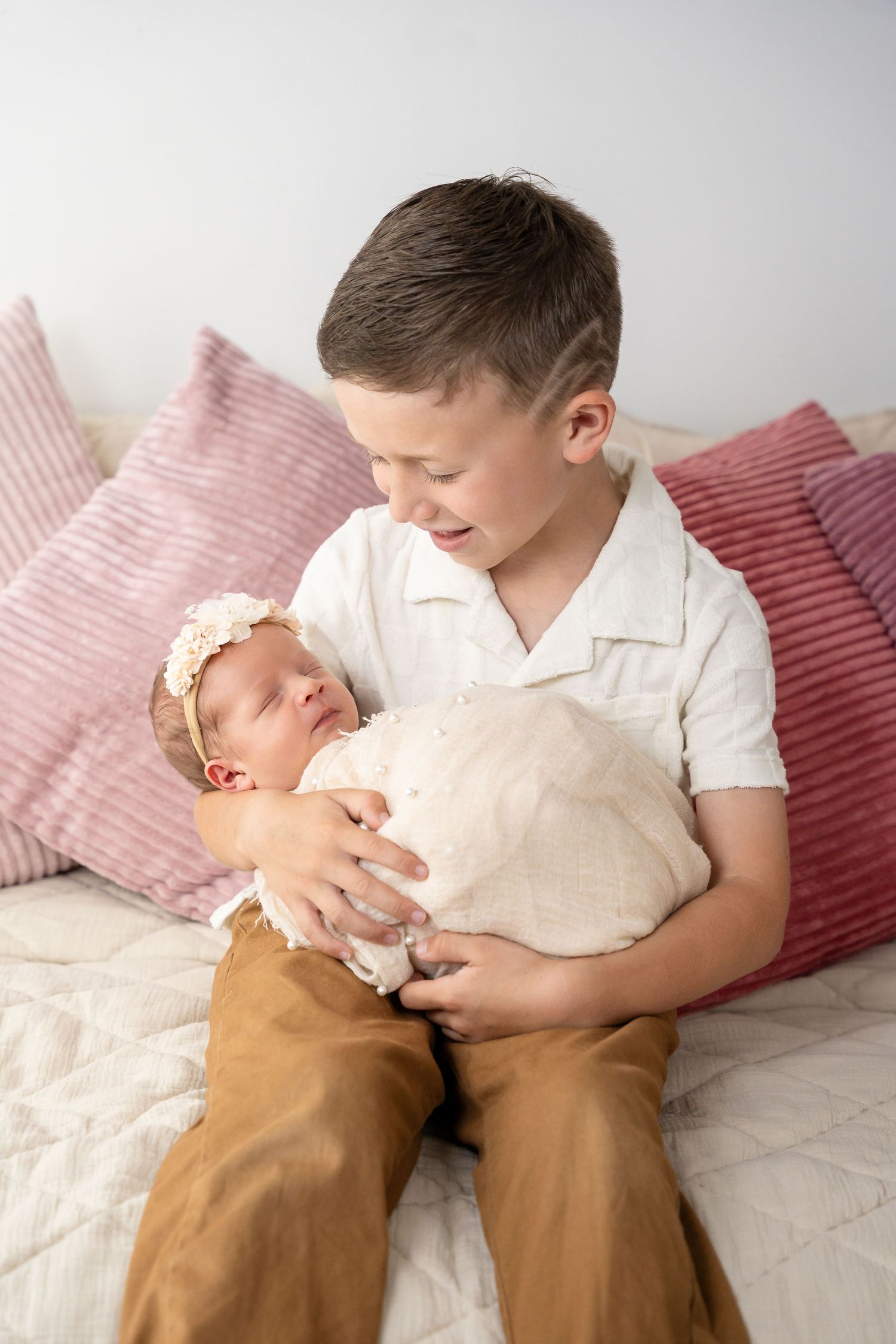 Siblings bonding moment as older child cradles newborn on white bedding with pink pillows.