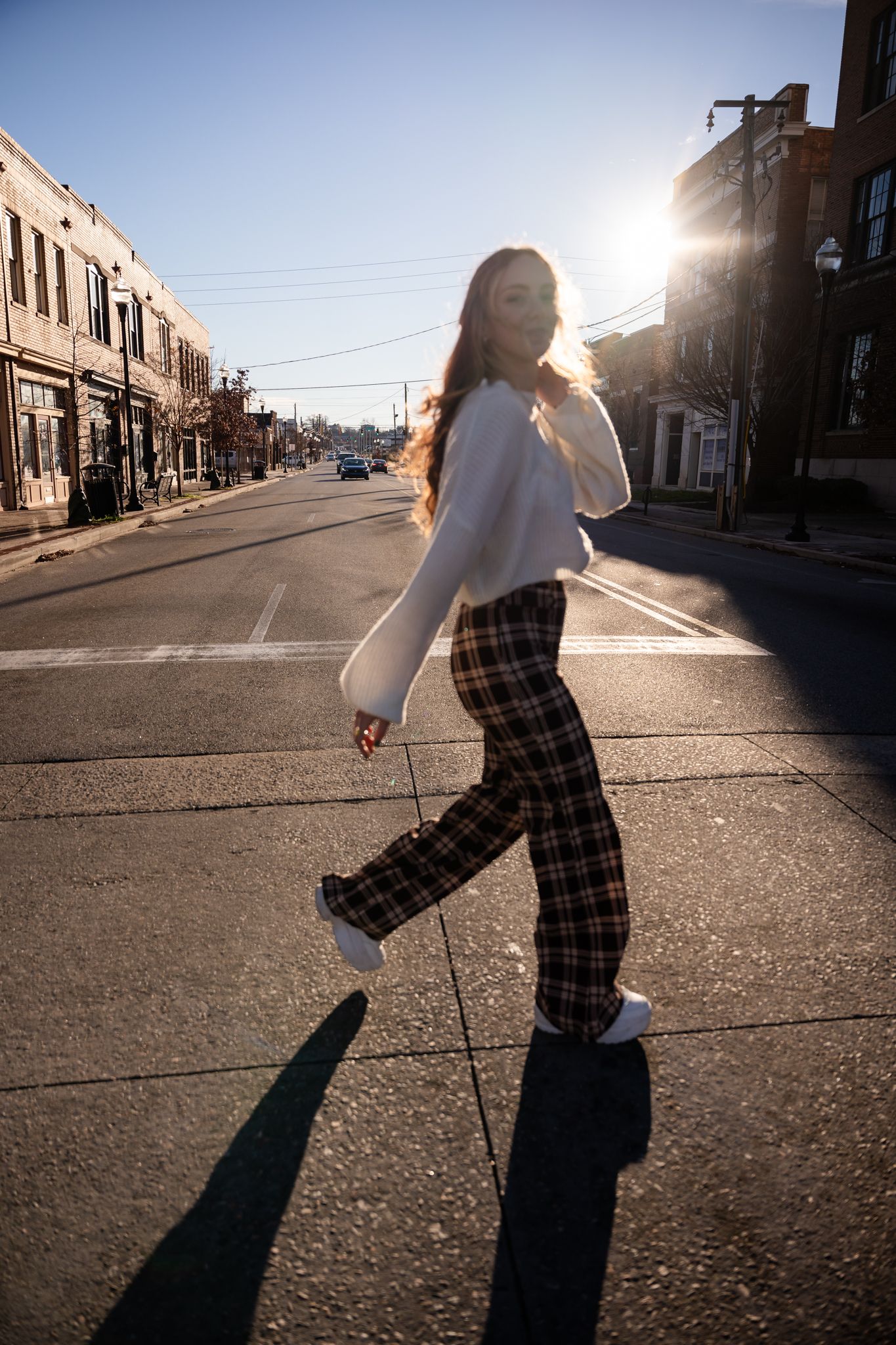 Dynamic street photography with long shadows featuring person in white sweater and plaid pants.