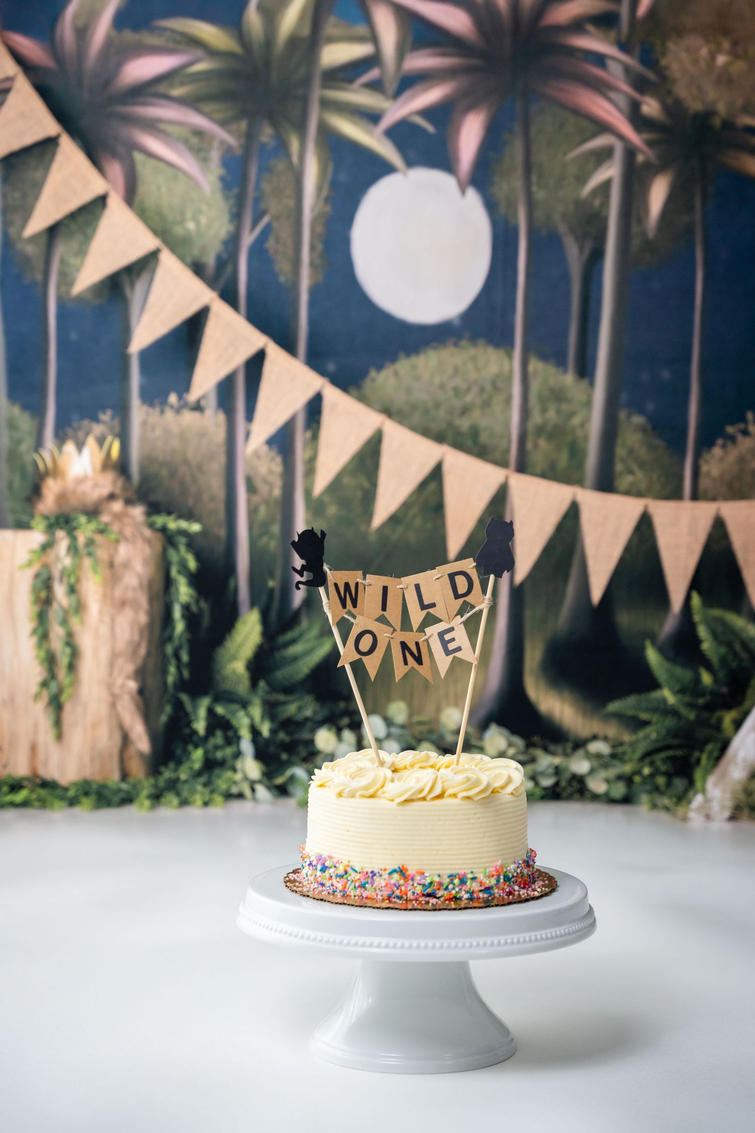 Four birthday cakes on white pedestals with bunting decorations against a dark background with greenery.