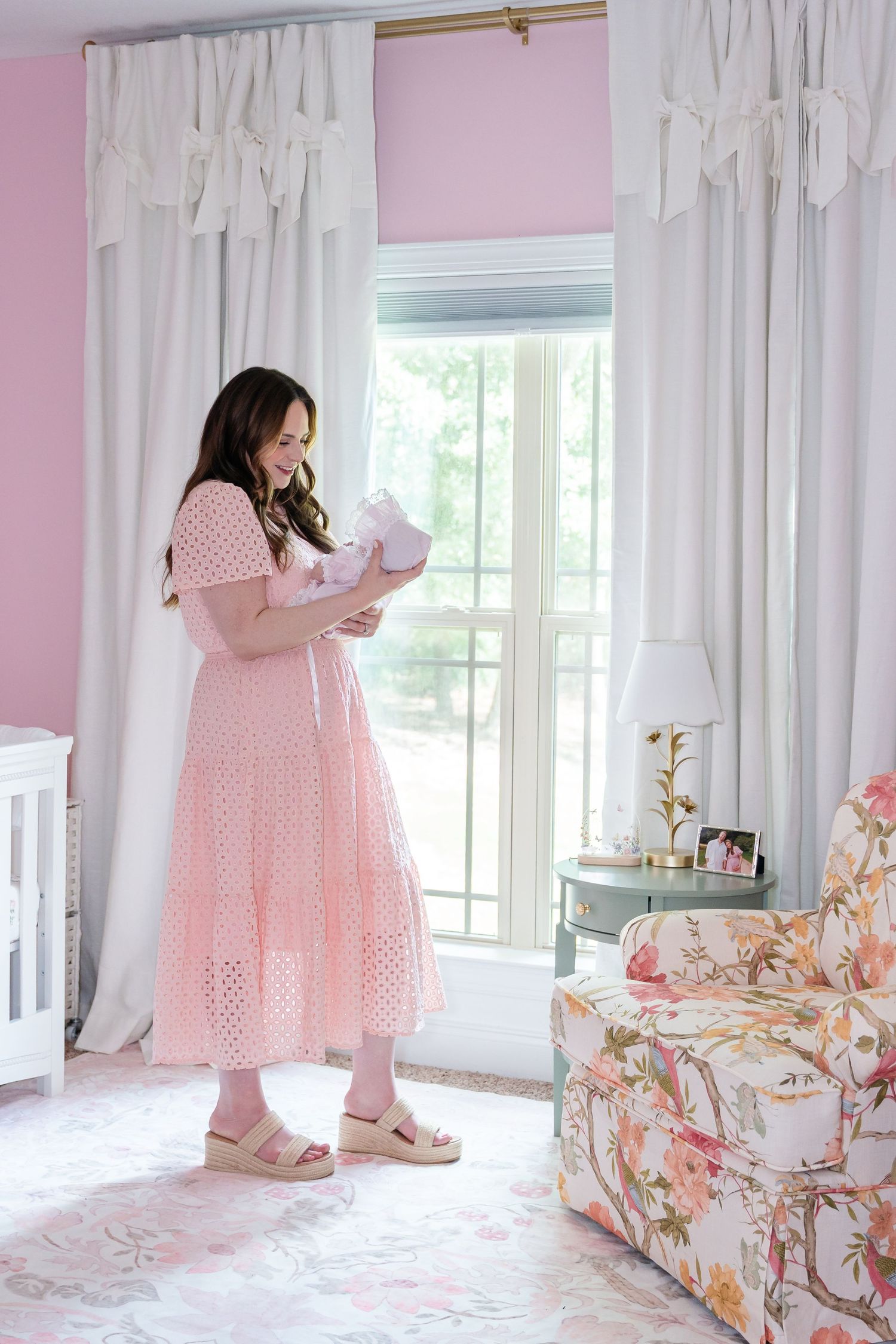 Woman in a pink eyelet dress holds a newborn in a pink nursery with white curtains, a floral armchair and a side table lamp.