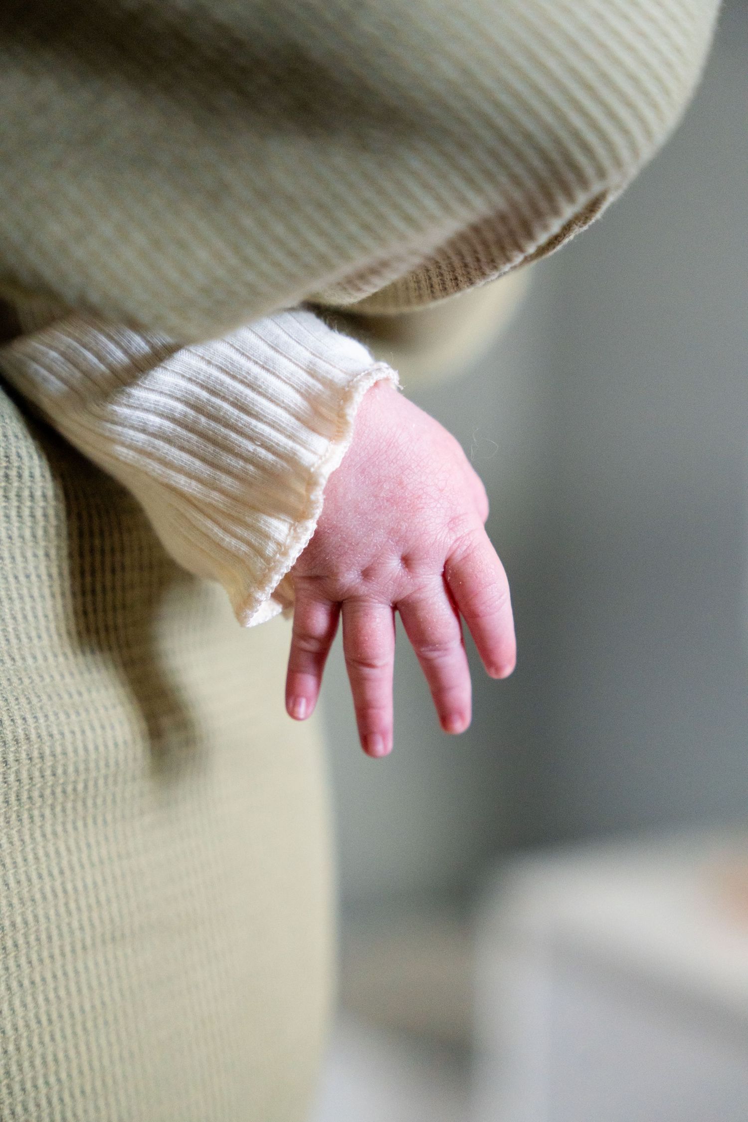 Close-up of a newborn baby's tiny pink hand peeking out from a soft white and beige knit sleeve.