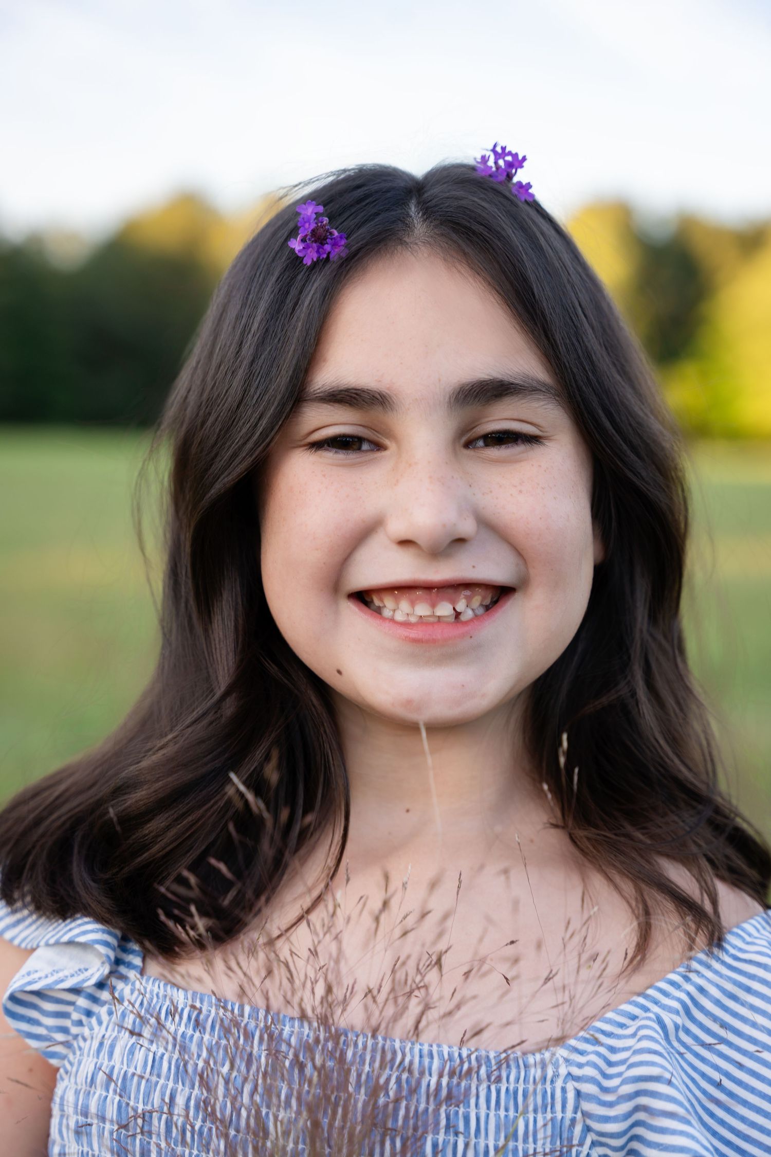 Smiling young girl with dark hair and purple flower clips wearing a blue striped top outdoors.