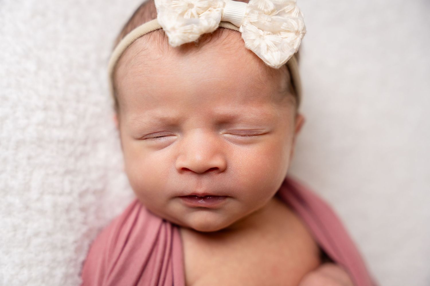 Sleeping baby with cream bow headband on textured white backdrop.