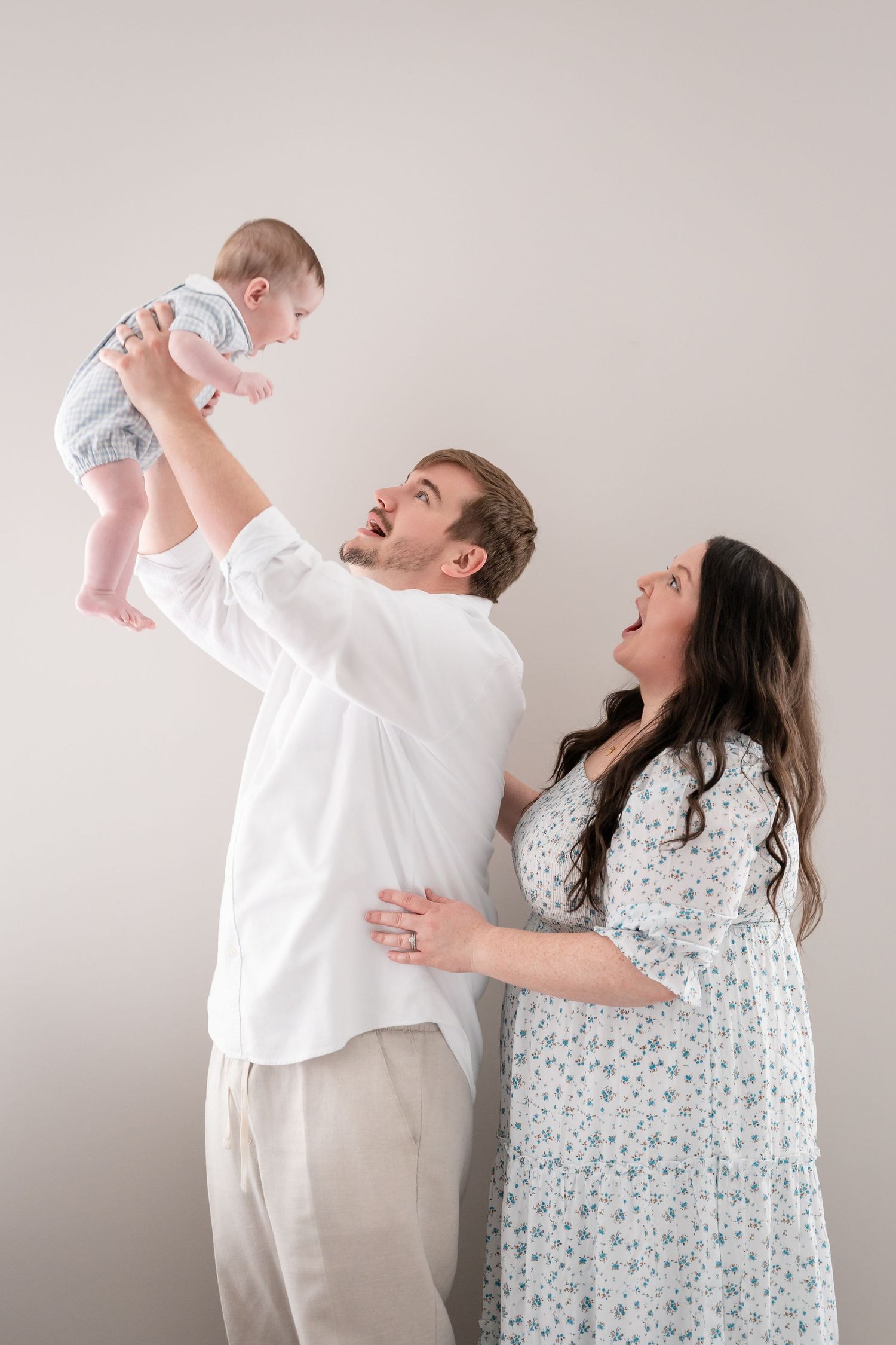 Parents playfully lift and interact with their infant while wearing white and floral outfits.