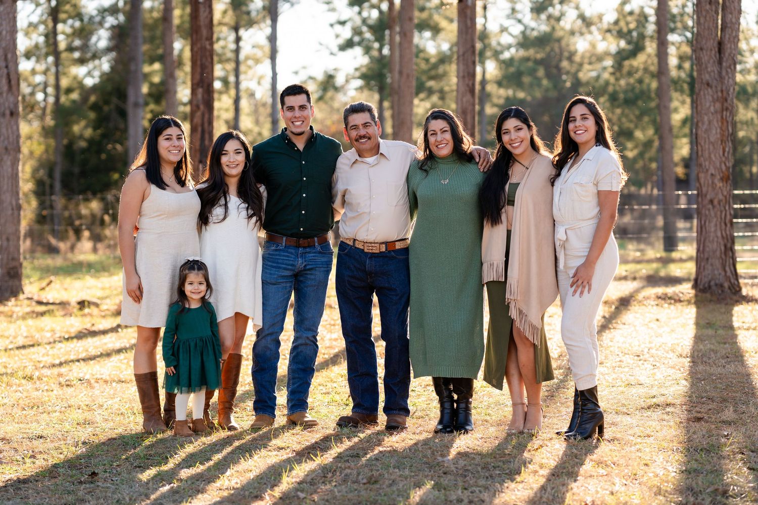A family group poses together in front of pine trees at sunset wearing coordinating white, green and neutral outfits.