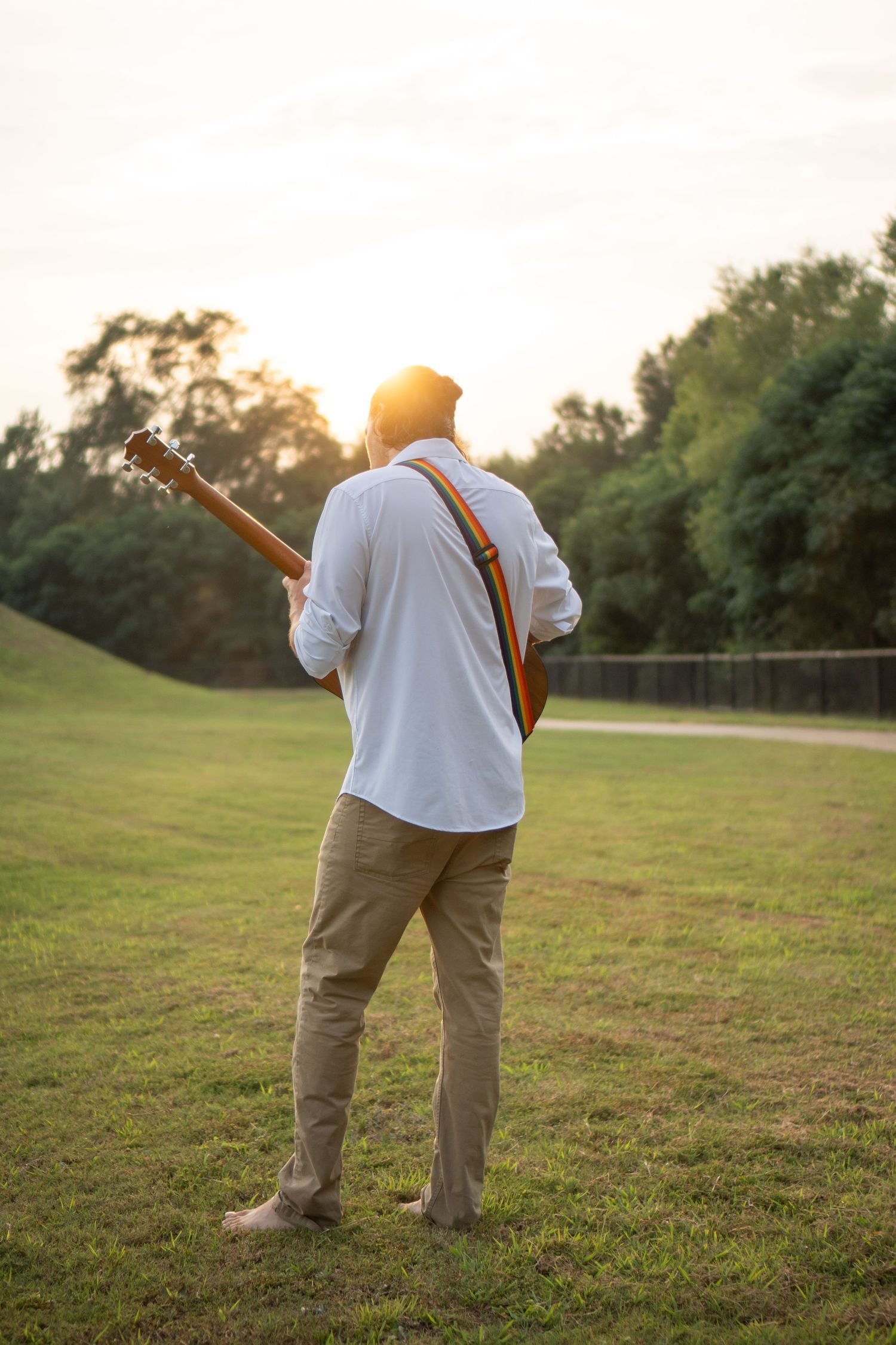Person with guitar standing in grassy field during golden hour sunset.