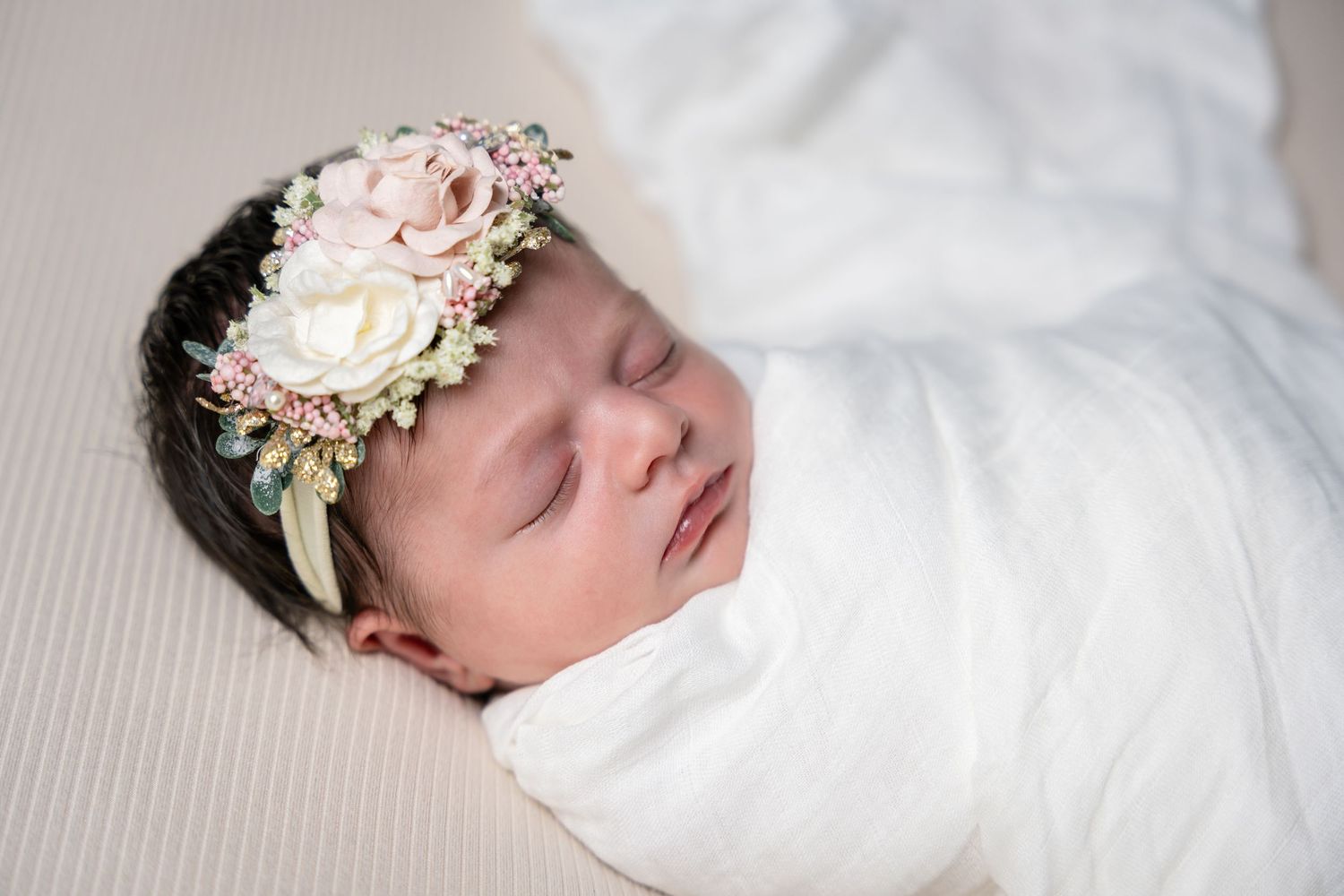 A sleeping newborn baby wearing a delicate pink floral headband rests peacefully on white bedding.