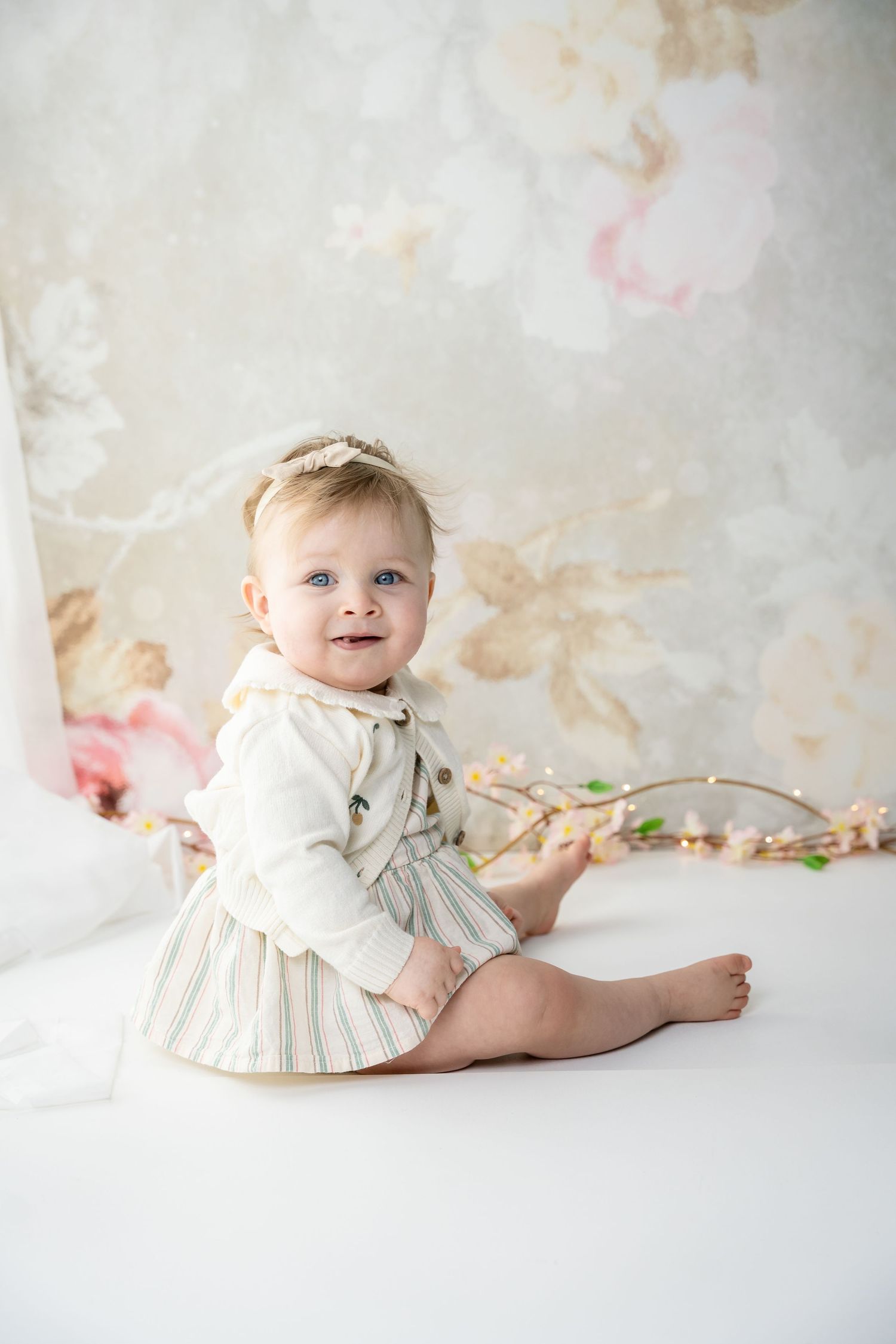 A baby in a cream-colored dress lies on a white backdrop with soft, dreamy lighting and floral accents.
