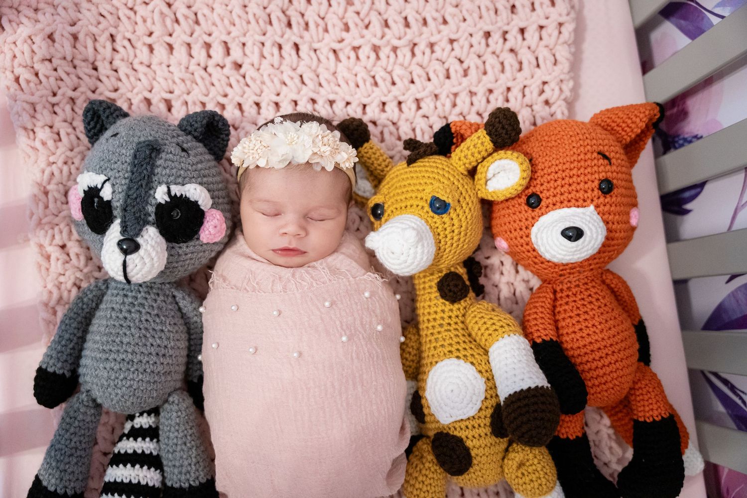 Sleeping newborn baby surrounded by crocheted stuffed animal toys including a raccoon, giraffe, and fox on a pink blanket.