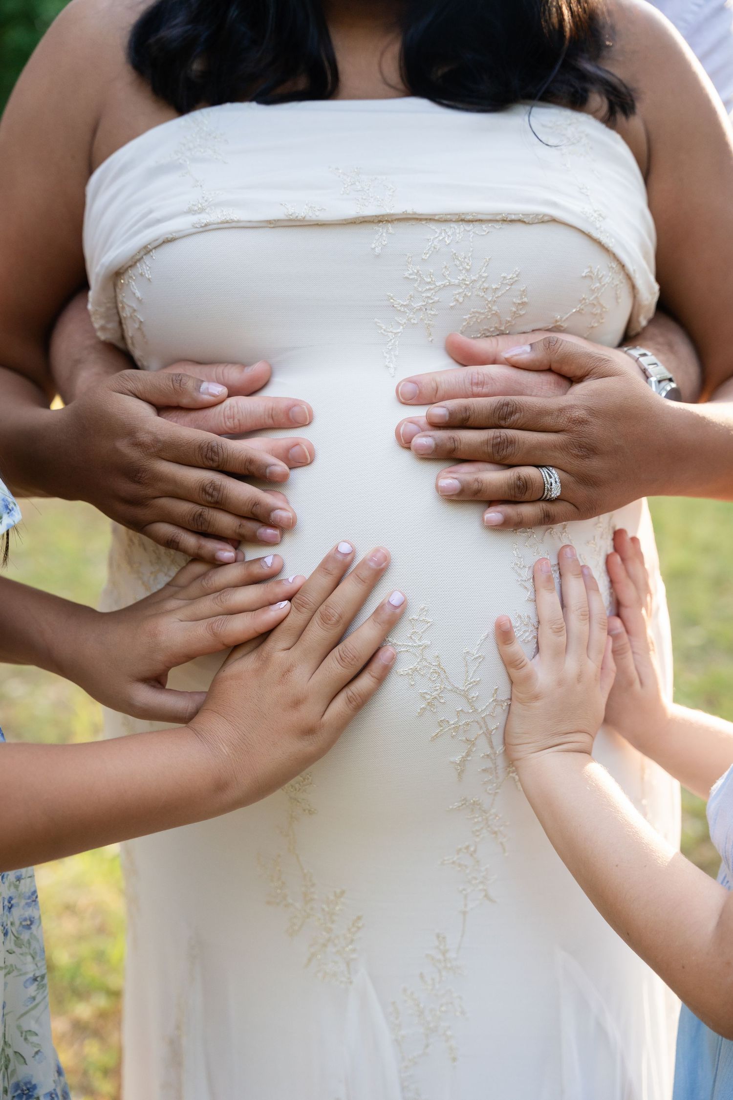 Multiple hands lovingly placed on a pregnant woman's belly in a white lace maternity gown.