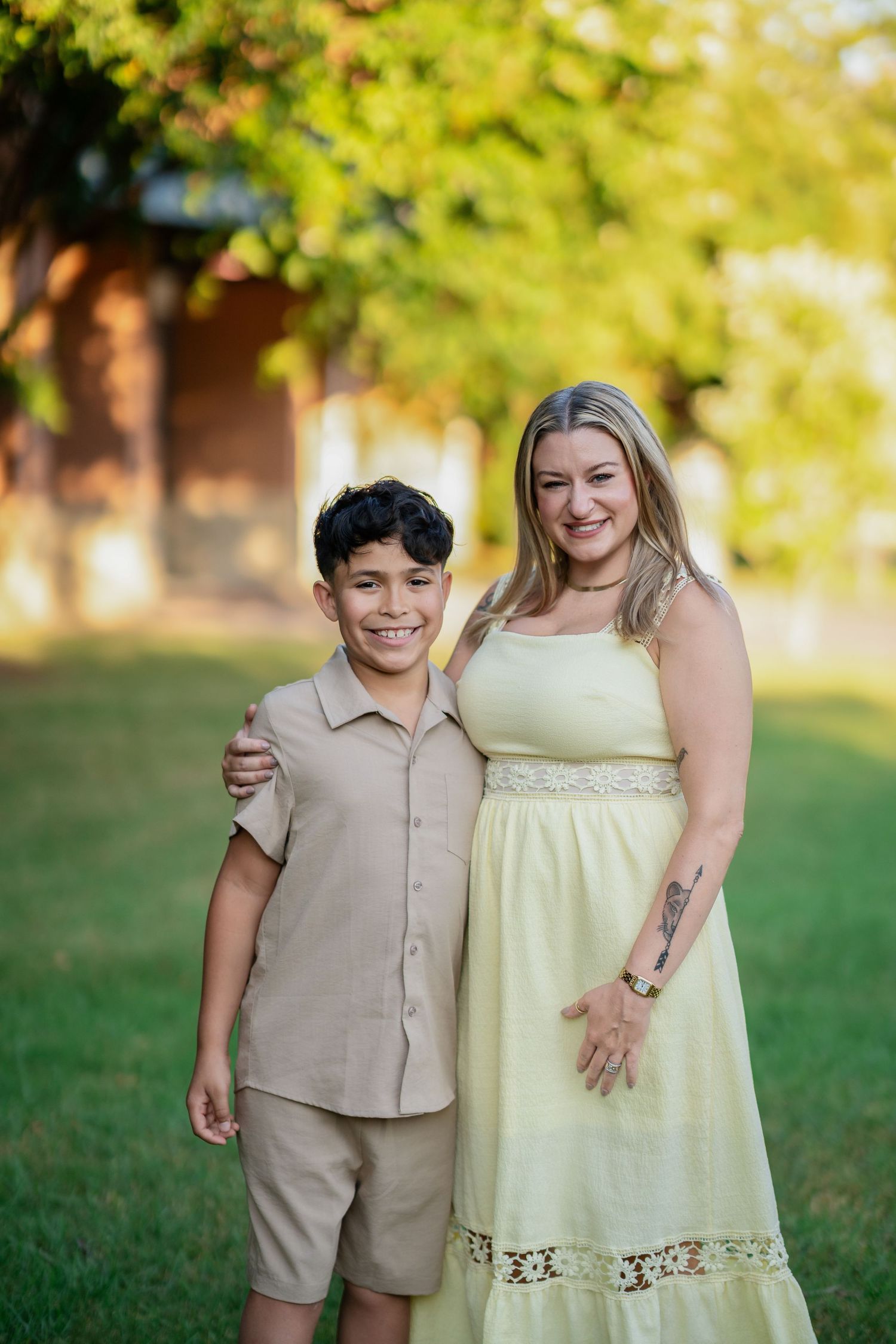 A woman in a yellow dress and a boy in tan outfit smile together outdoors in a green park setting.