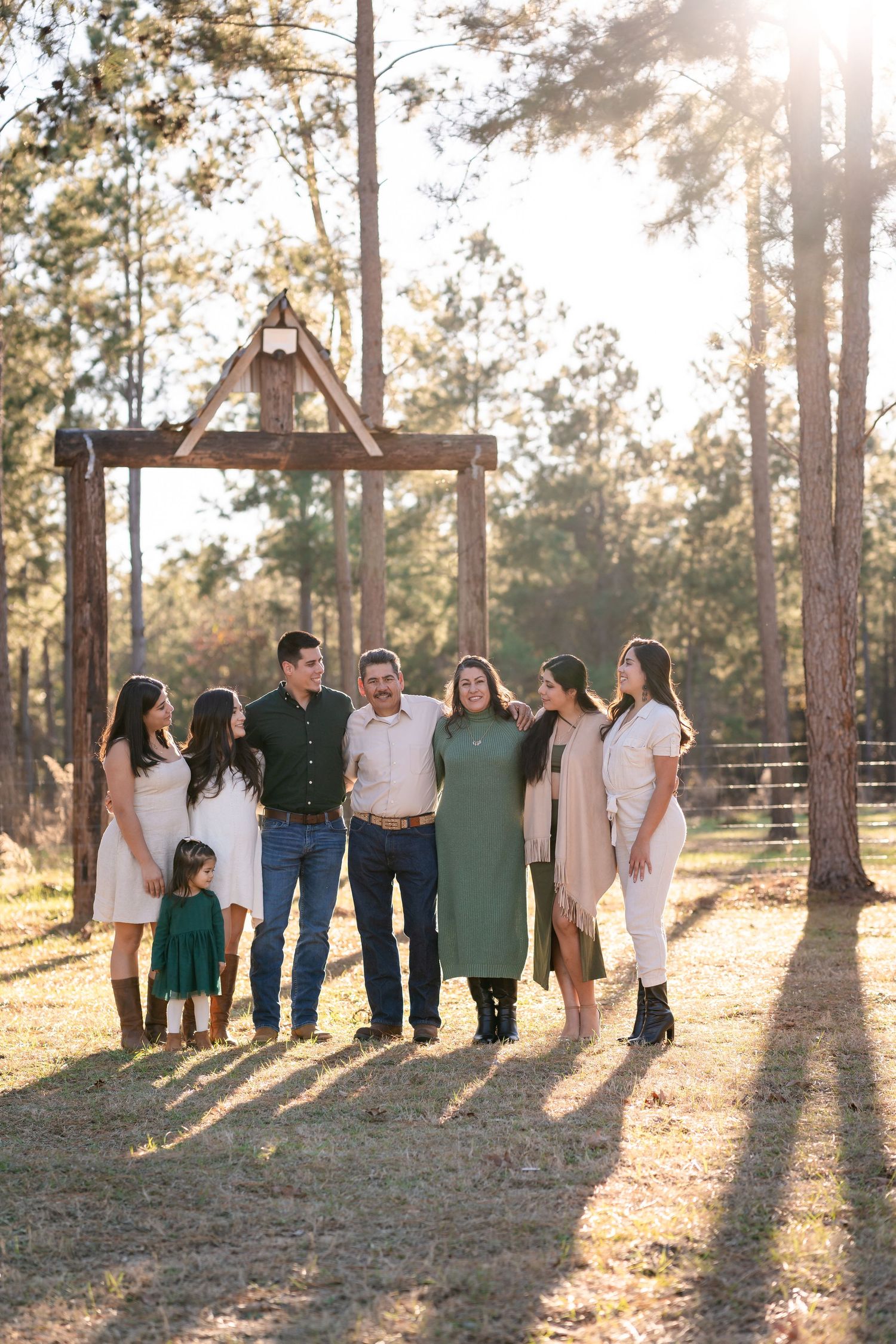 Family gathering beneath wooden archway with long shadows cast by evening sunlight through tall pine trees.