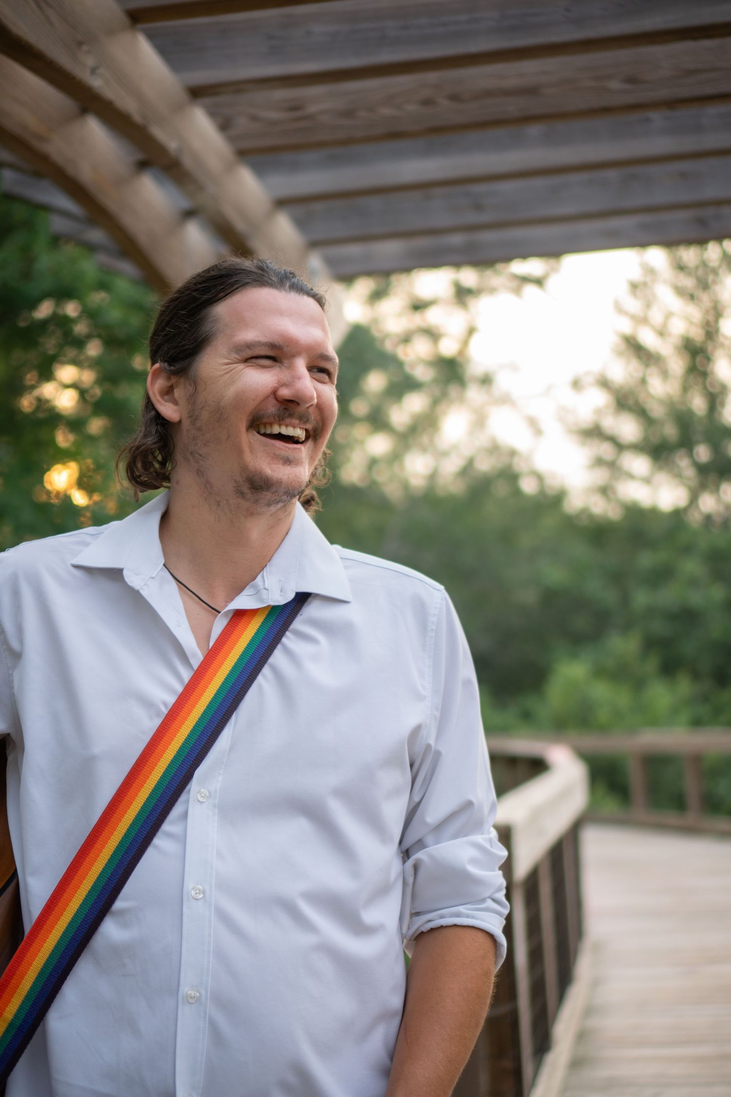 Musician wearing white shirt with rainbow stripe sash on wooden deck with trees in background.