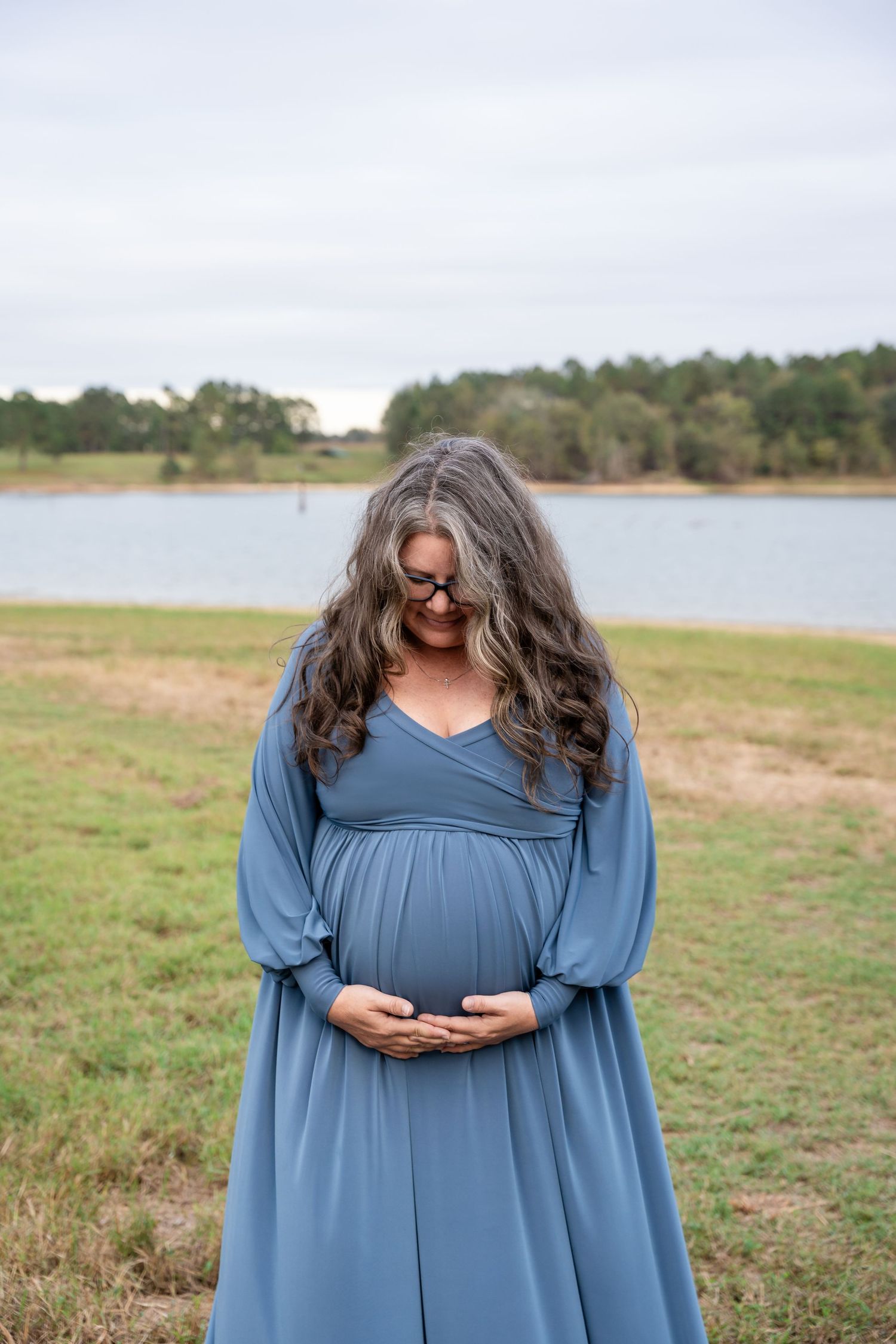 Person in flowing blue dress with long hair stands in grassy field by lake on cloudy day.