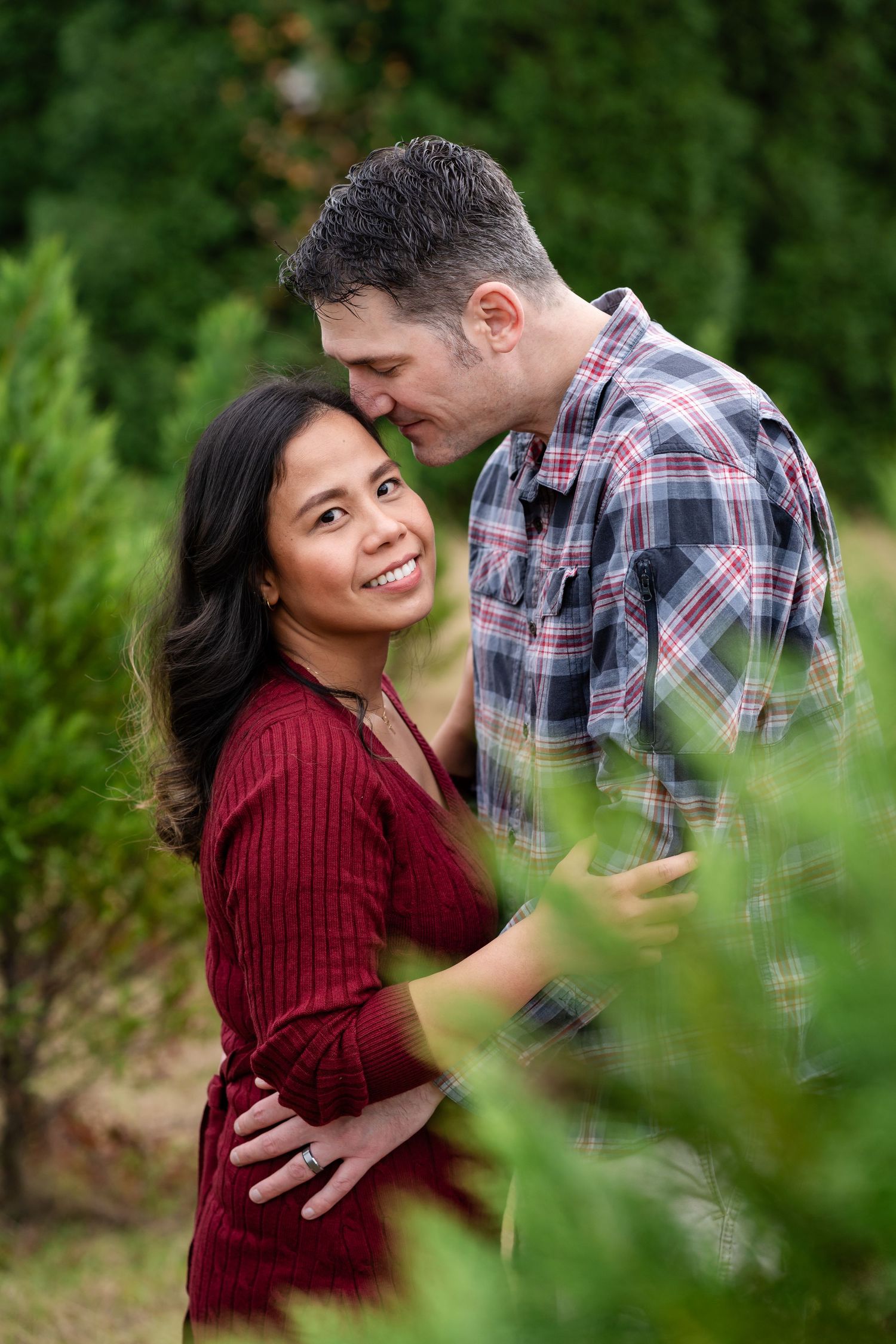 A couple embraces in a romantic outdoor photoshoot against green foliage in burgundy and plaid.