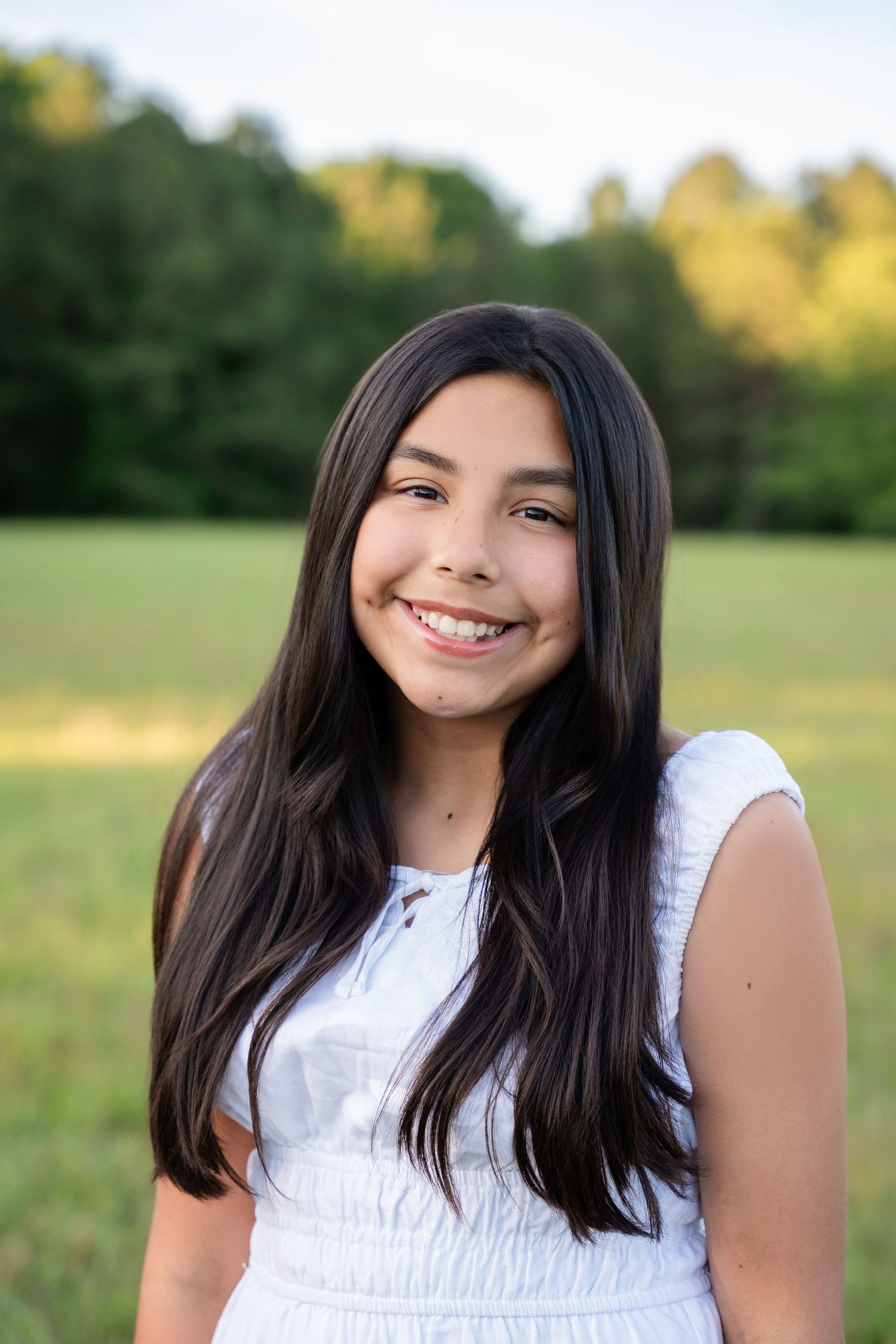 Smiling teenage girl with long dark hair wearing a white dress outdoors in a green field.