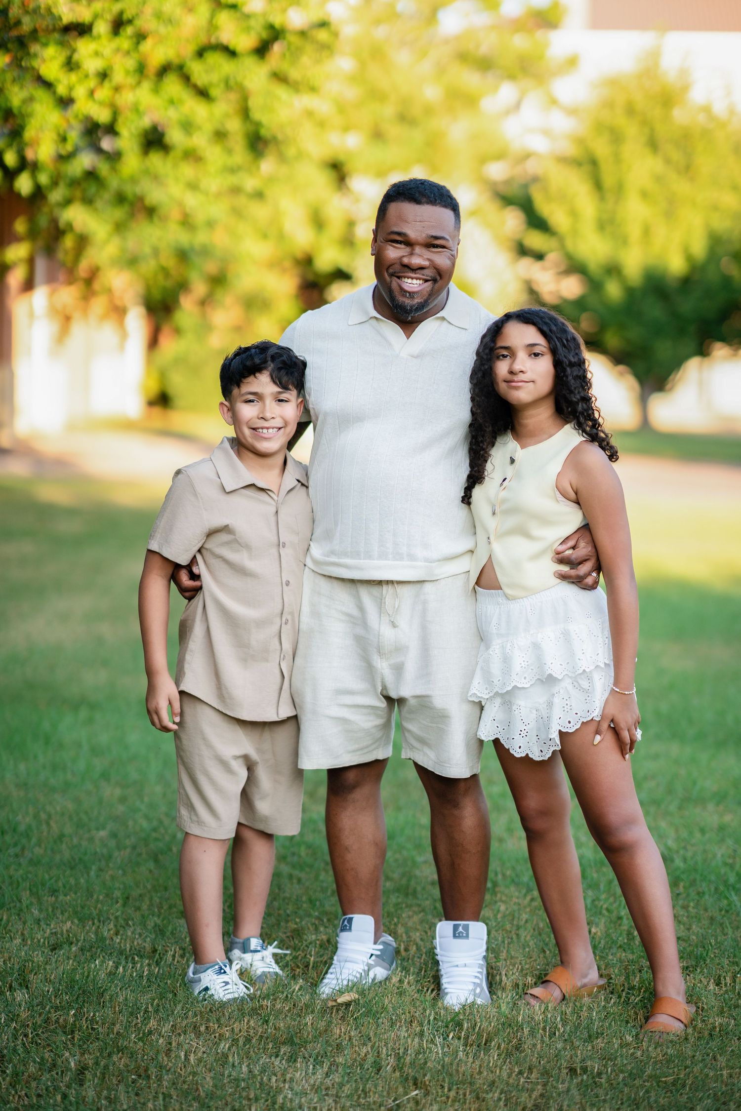A smiling man poses outdoors on green grass with two children, all wearing neutral-toned outfits in a sunny park.