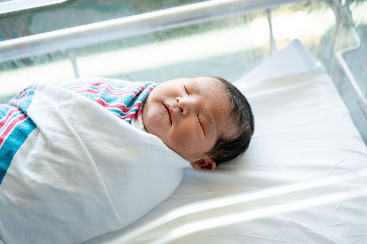 Sleeping newborn baby swaddled in a white and striped hospital blanket, resting peacefully in a clear bassinet.