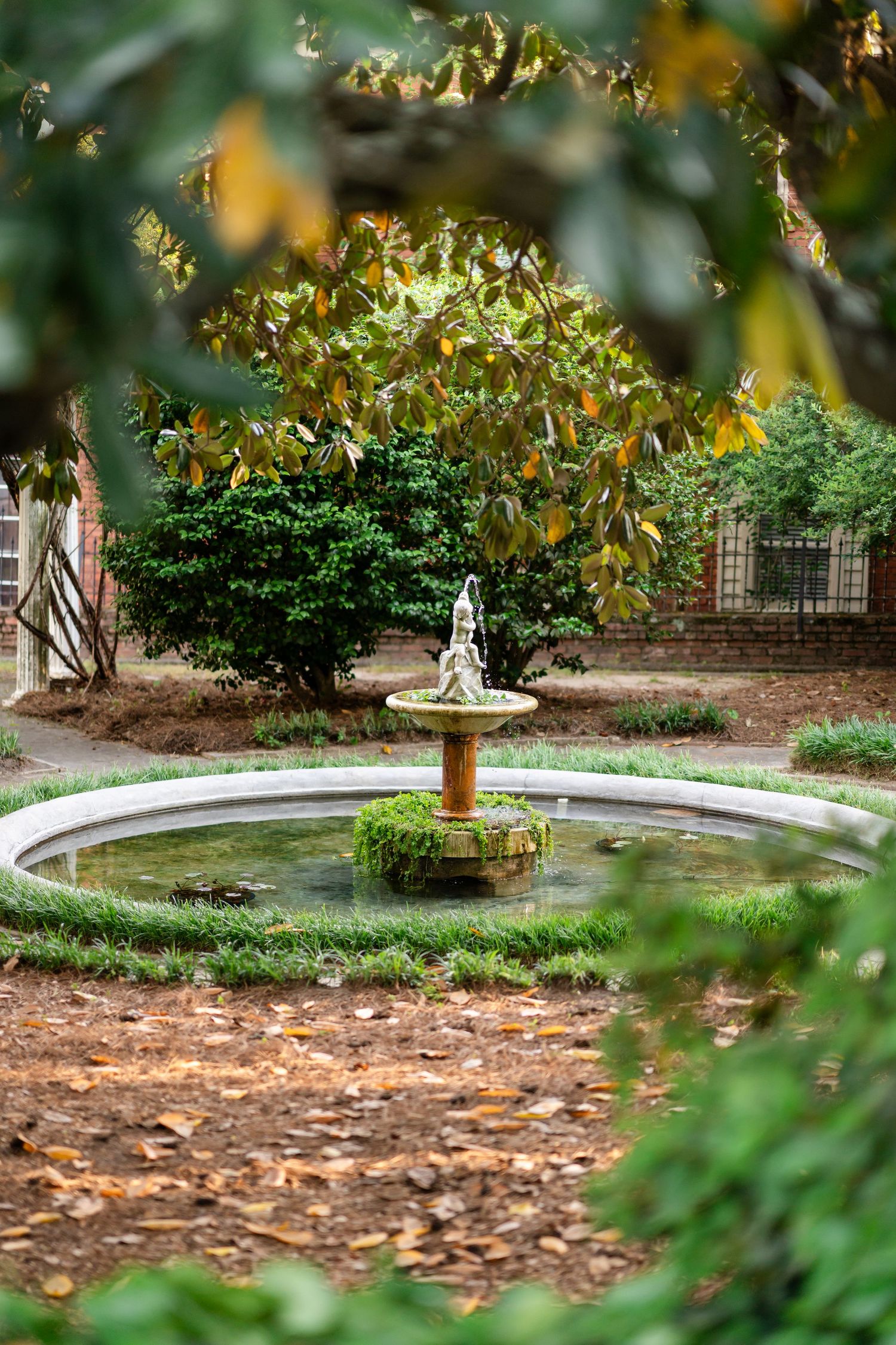 Decorative garden fountain surrounded by lush greenery and fallen autumn leaves in a tranquil setting.