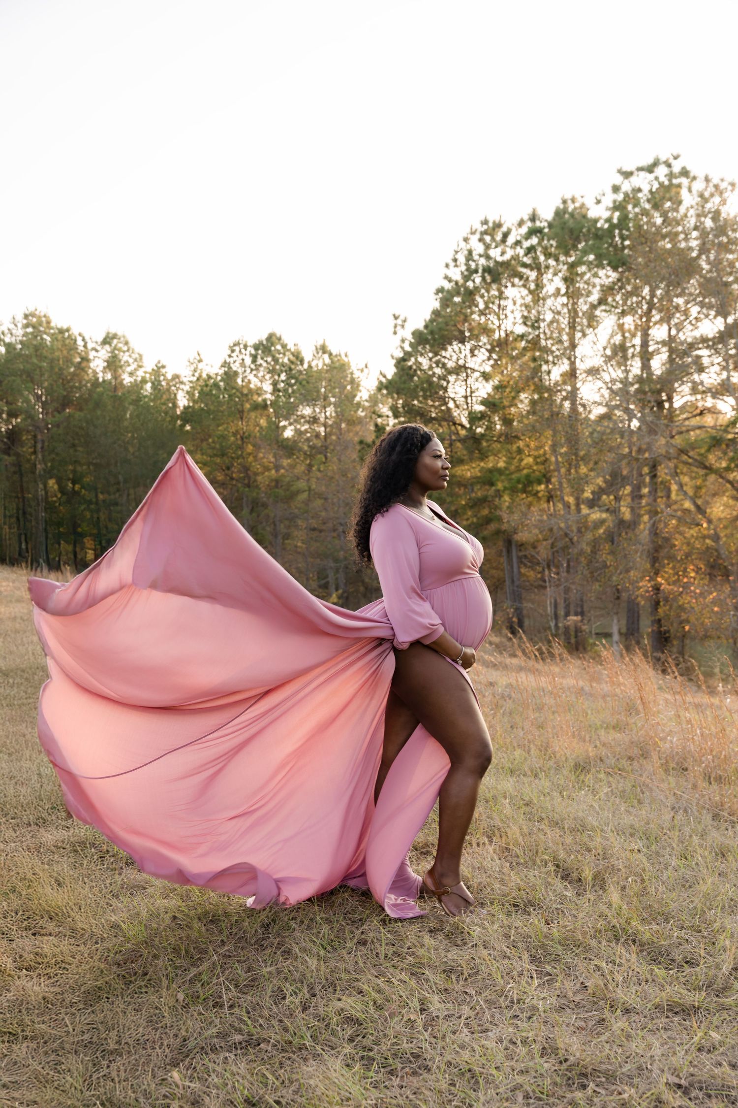 Pregnant woman in flowing pink maternity gown standing in a field with trees at golden hour.