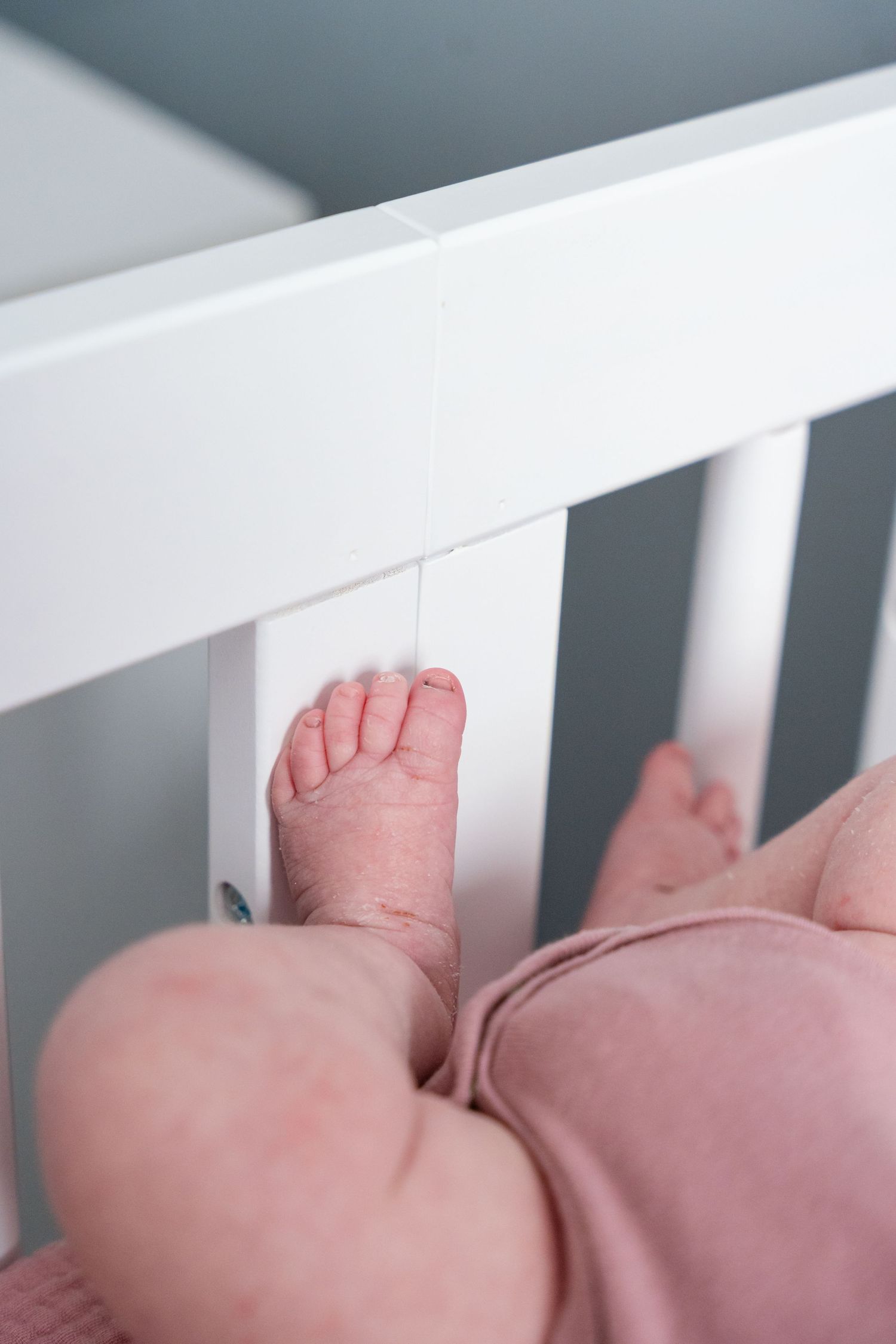 Close-up of tiny baby feet resting against white crib rails, with infant wearing pink outfit.