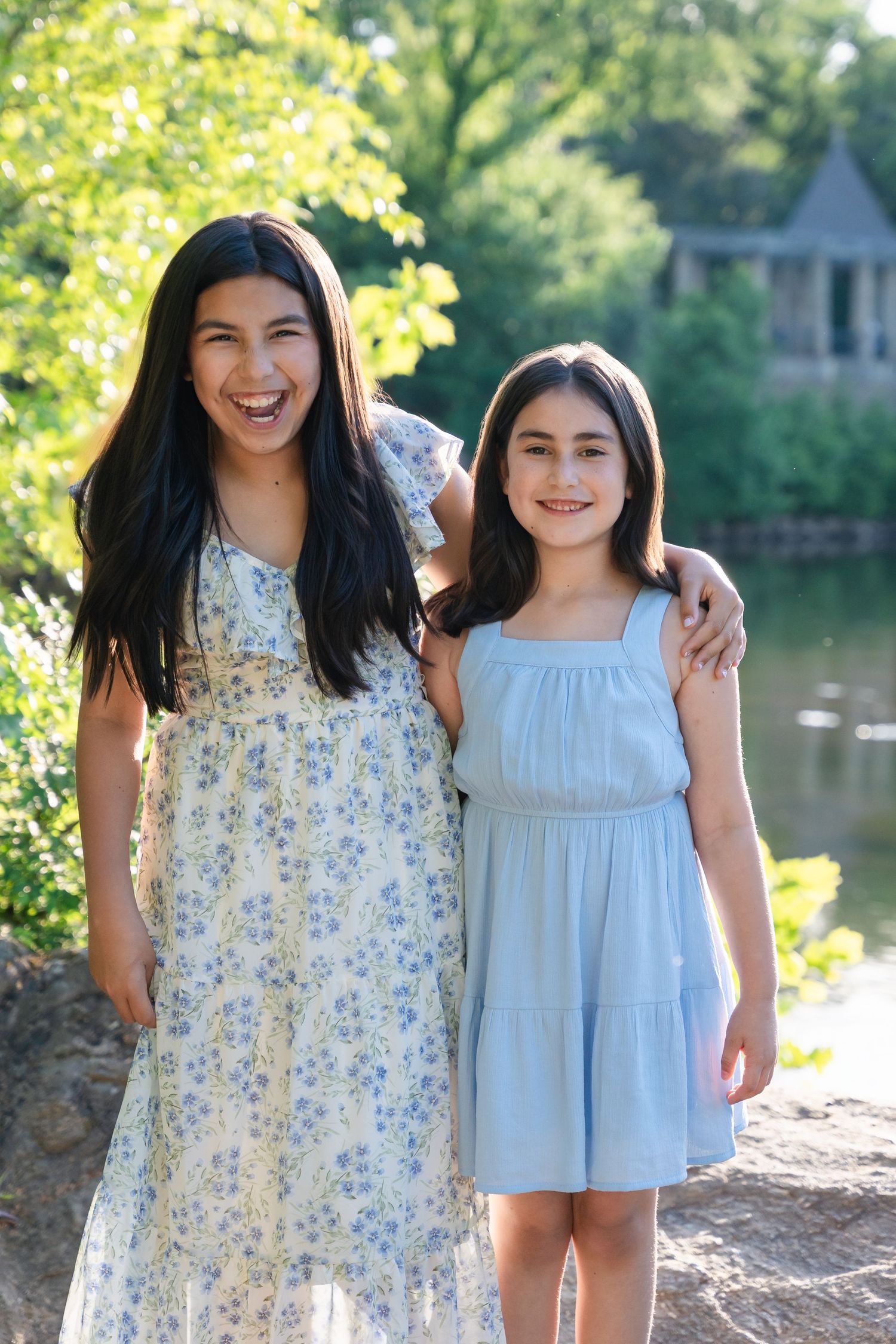 Two smiling sisters in floral and light blue dresses pose with arms around each other beside a calm lake on a sunny day.