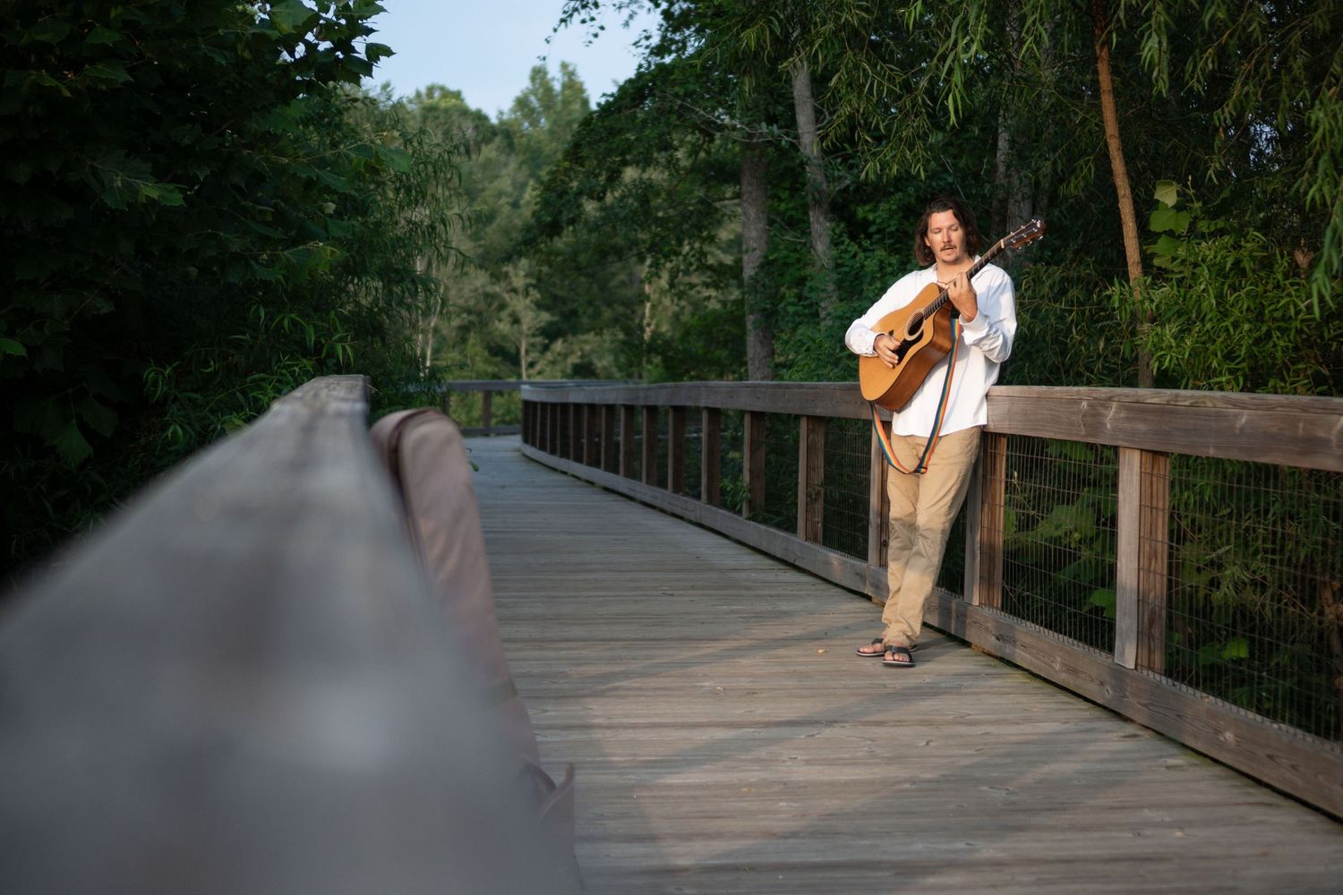 Distant view of musician playing guitar on long wooden bridge surrounded by trees.