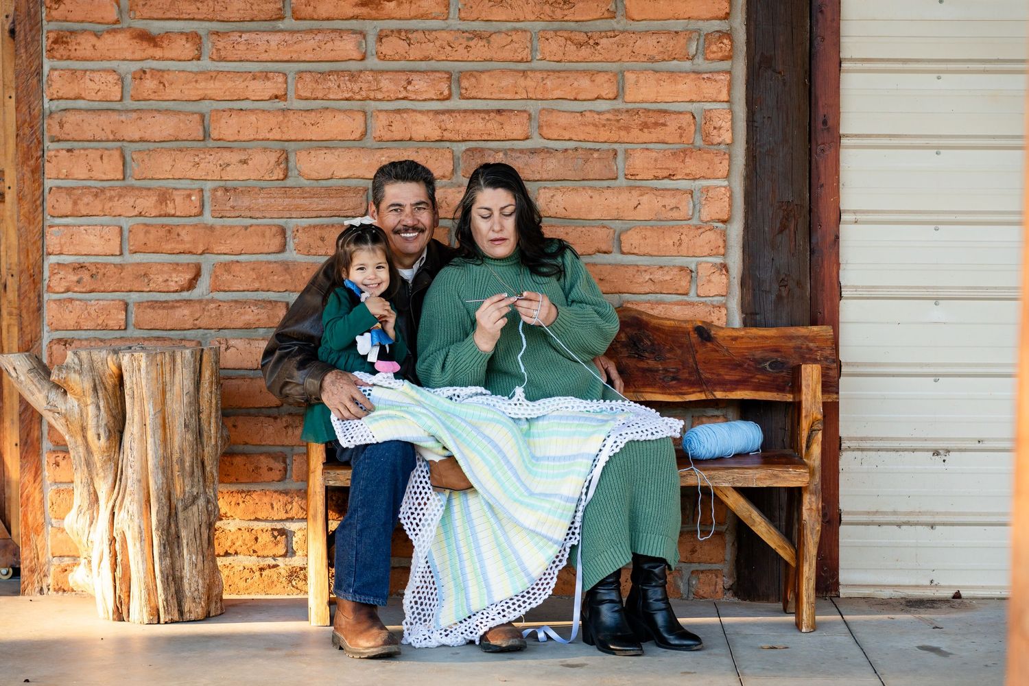 Three people sit on a wooden bench sharing a knitting moment on a rustic porch.