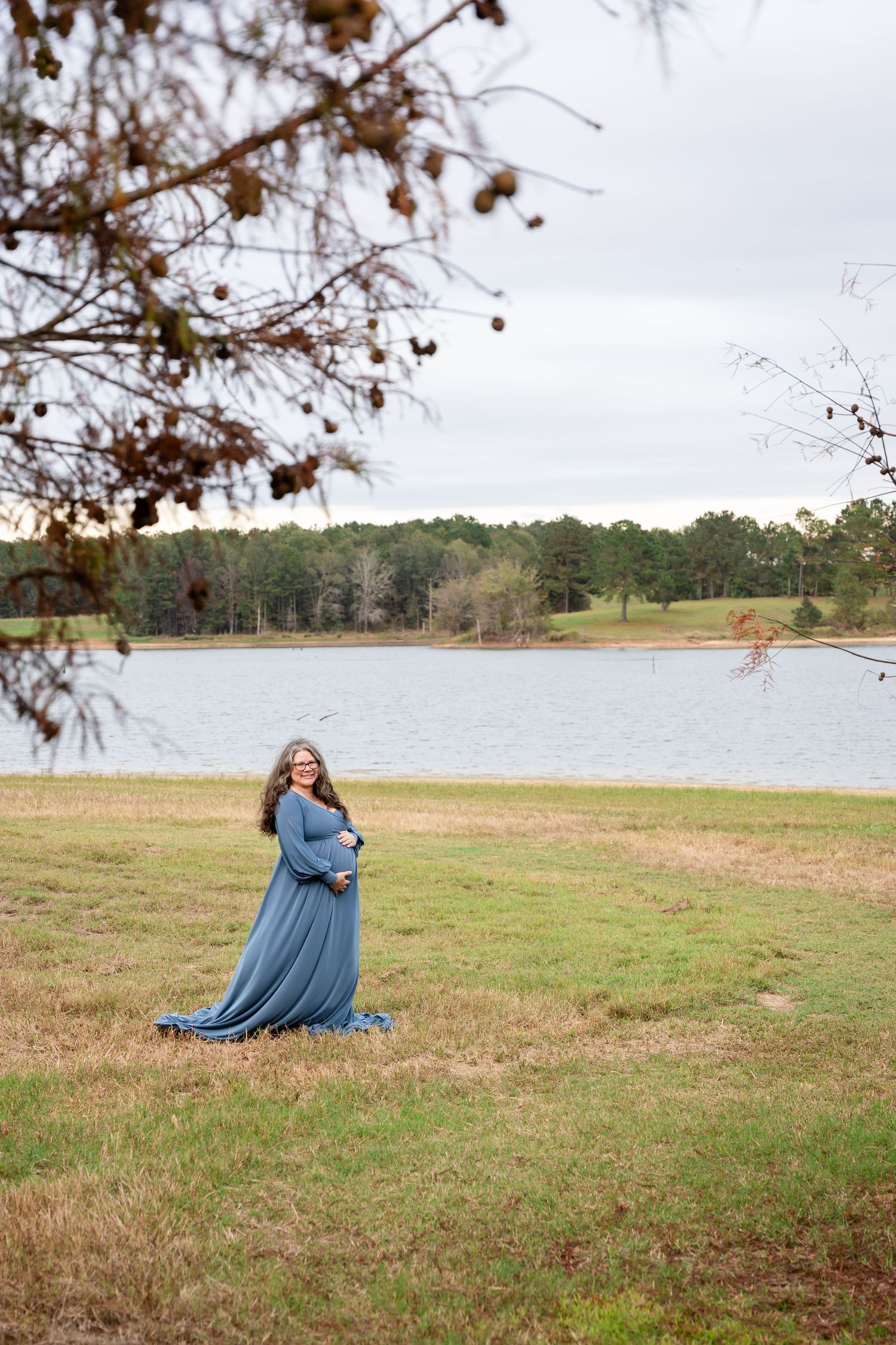 Person in flowing blue dress poses dramatically on grassy field near lake with autumn trees.