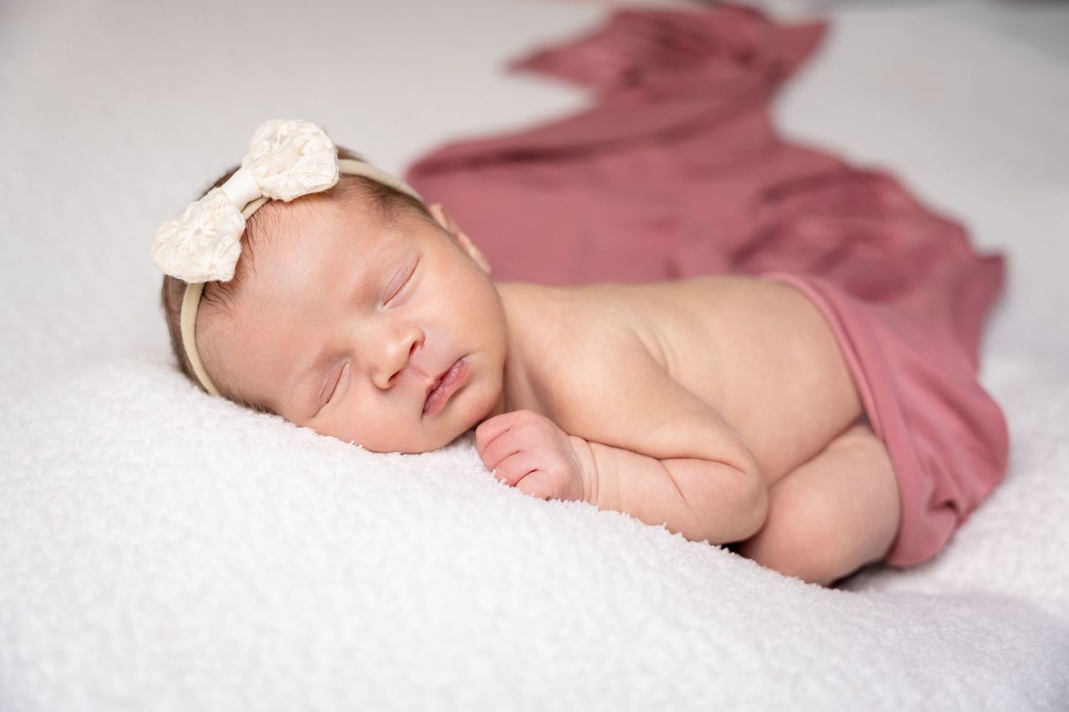 Newborn in dusty rose swaddle lying on white background.