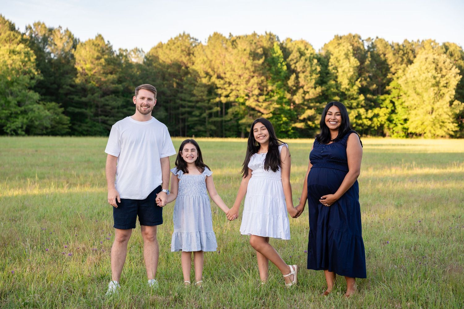 Smiling family of four holding hands in an open green field at golden hour.