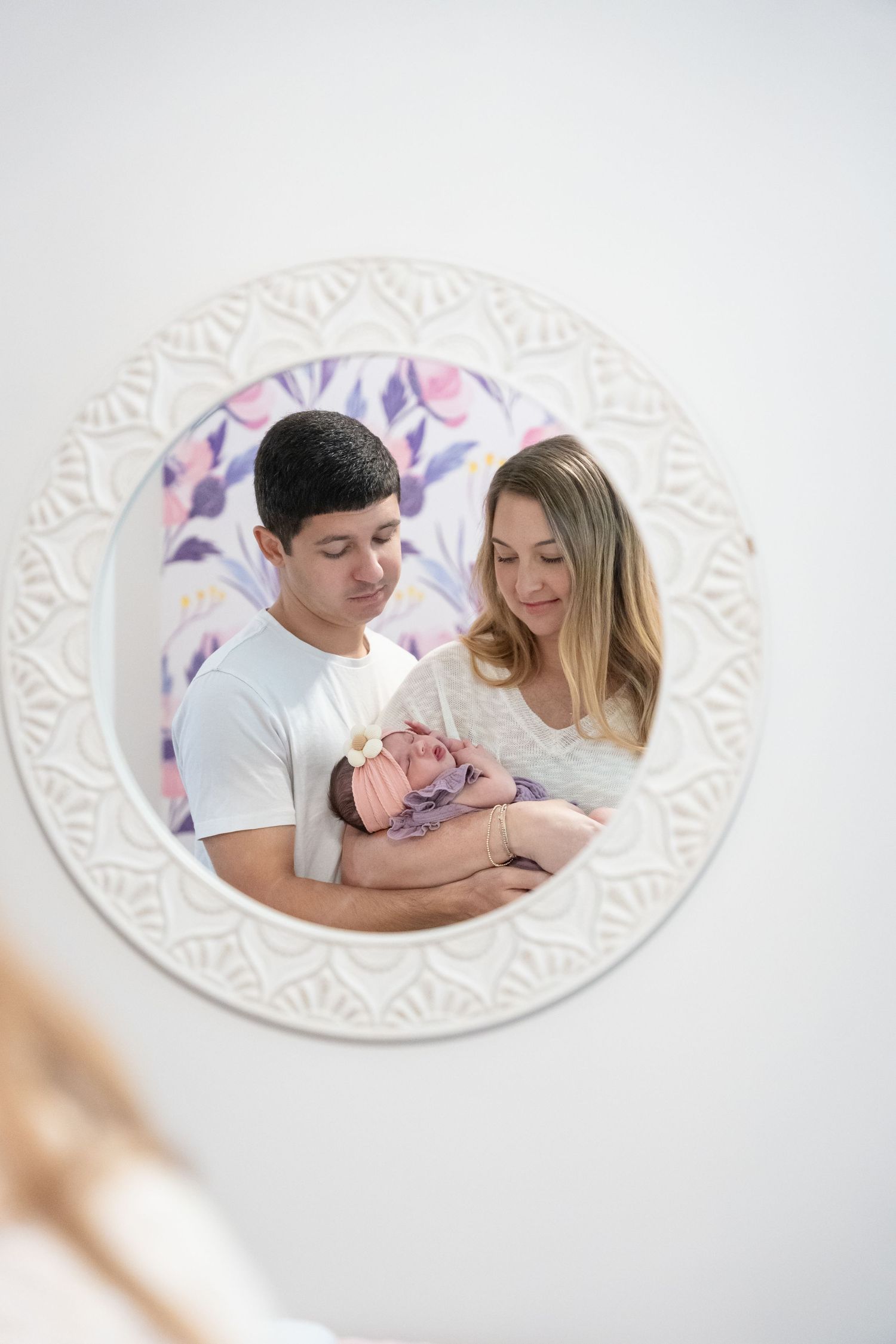 Young parents lovingly gaze at their newborn baby, reflected in a decorative round white mirror.