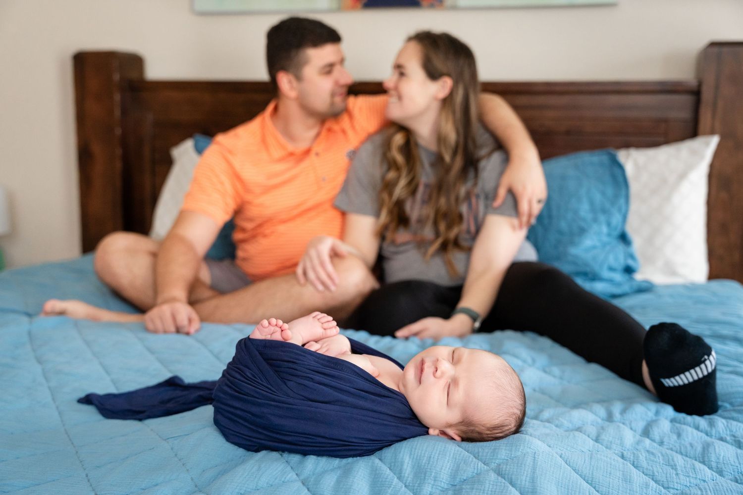 Family relaxing on bed with sleeping newborn in navy wrap.