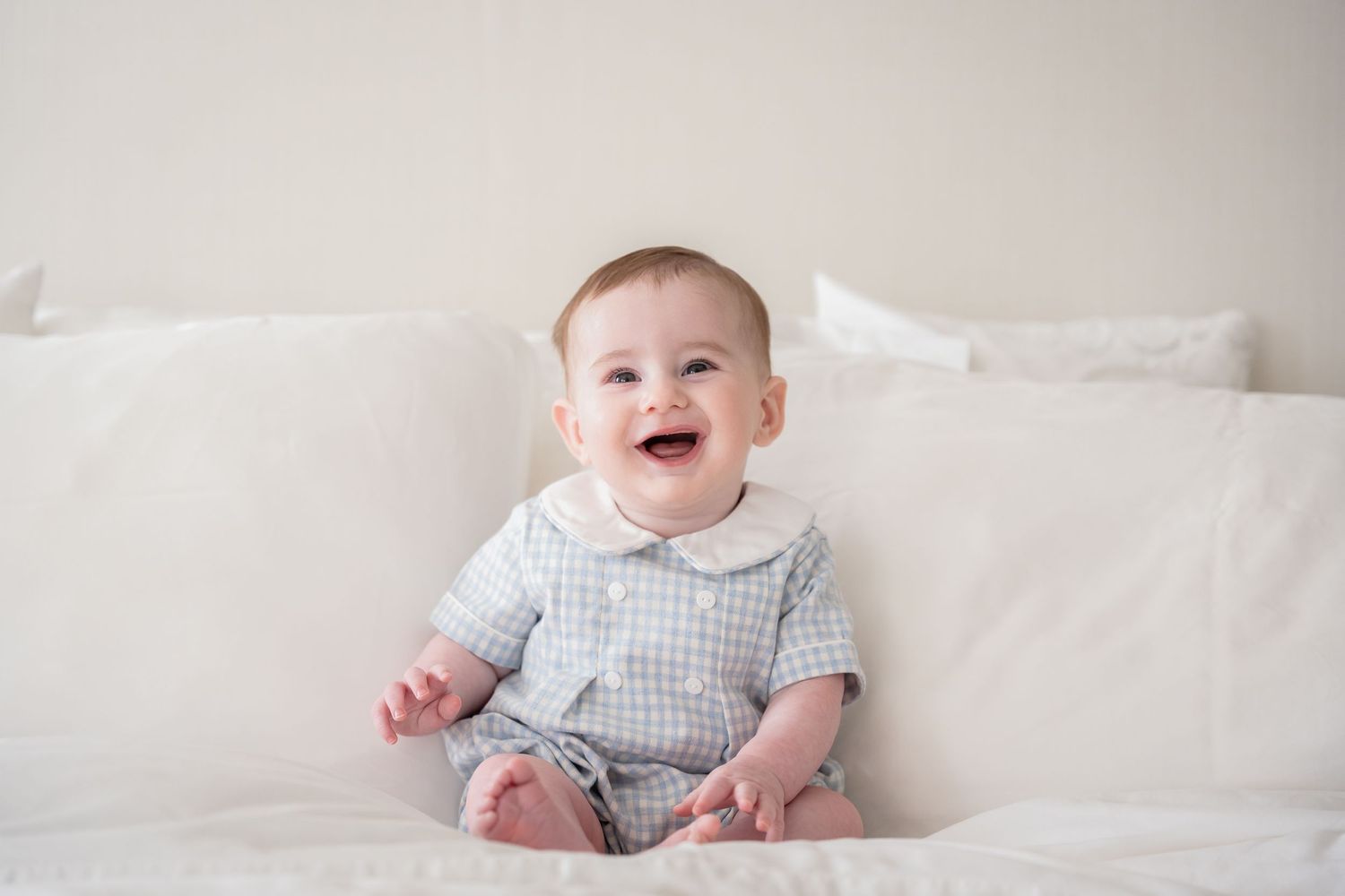 A baby in a checkered outfit sits on white bedding while smiling and laughing.