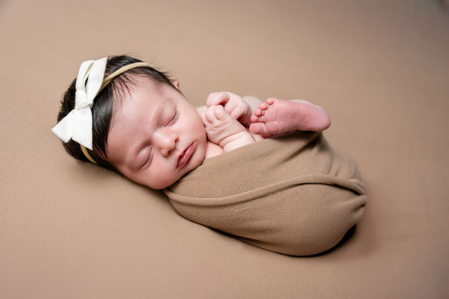 Sleeping newborn baby wrapped in brown swaddle with white bow headband lying on brown backdrop.