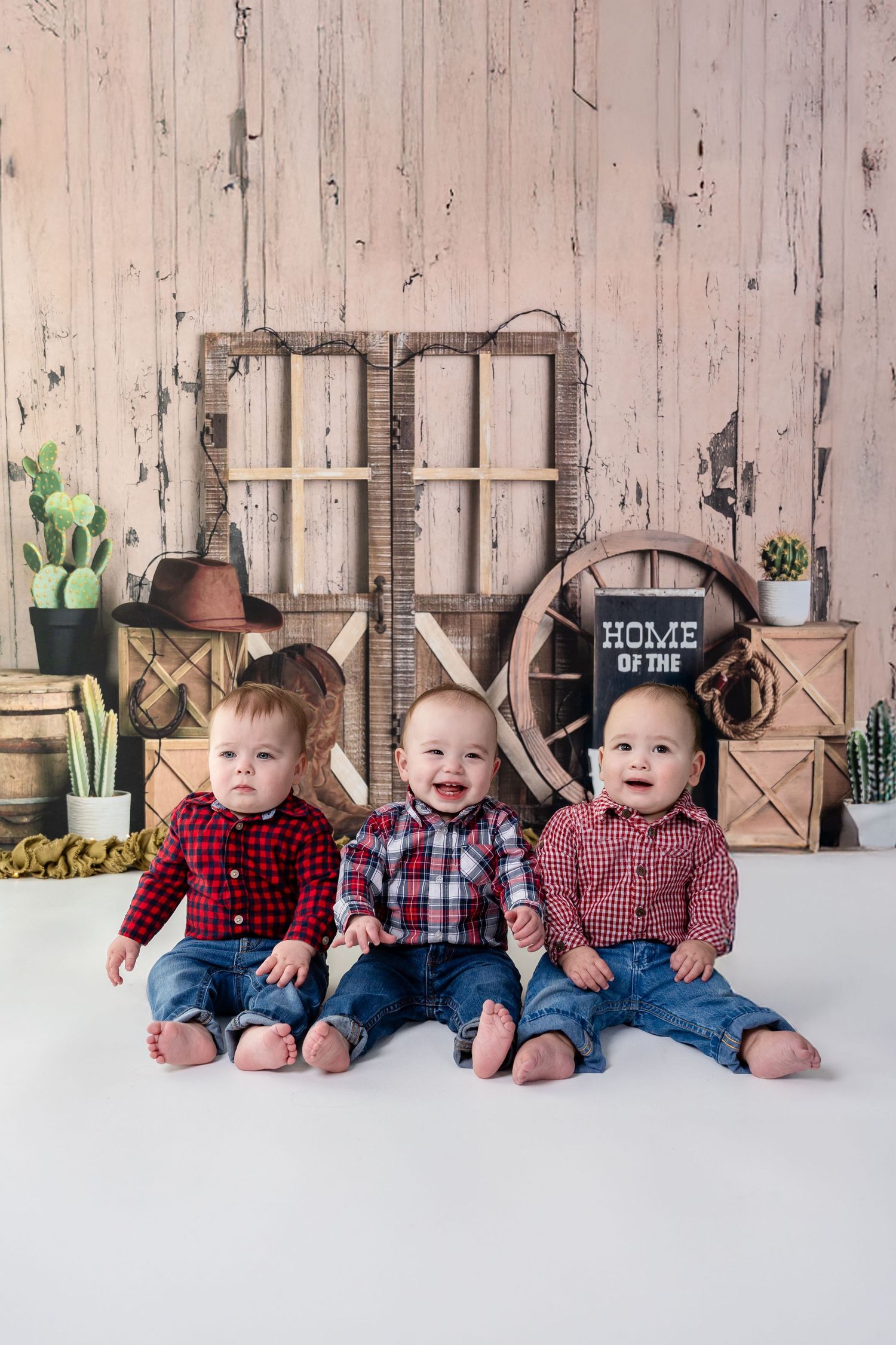 Three babies wearing plaid shirts and jeans sit together against a rustic farmhouse backdrop with cactus plants and home decor.