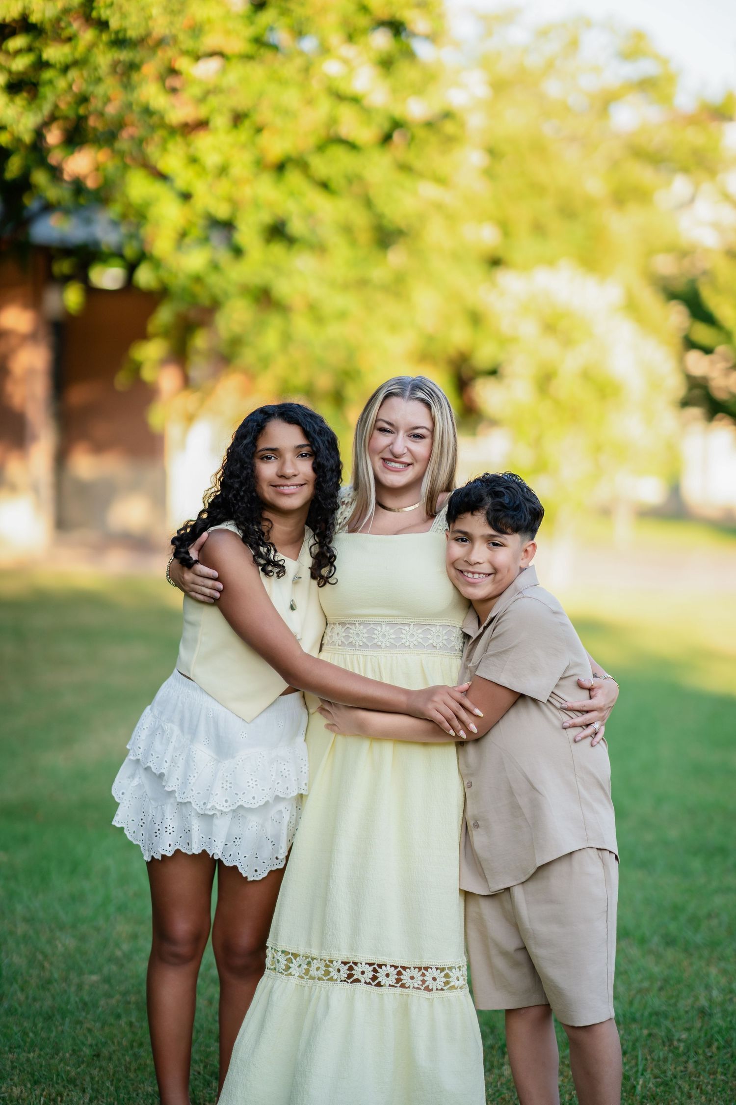 Blonde mother in yellow dress hugs two children outdoors on green grass in summer.