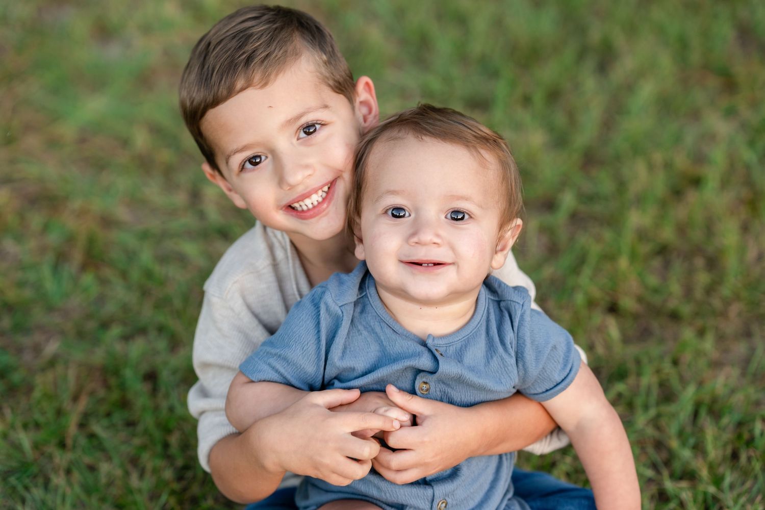 Two young brothers smiling outdoors, older boy hugging baby sibling on green grass background.