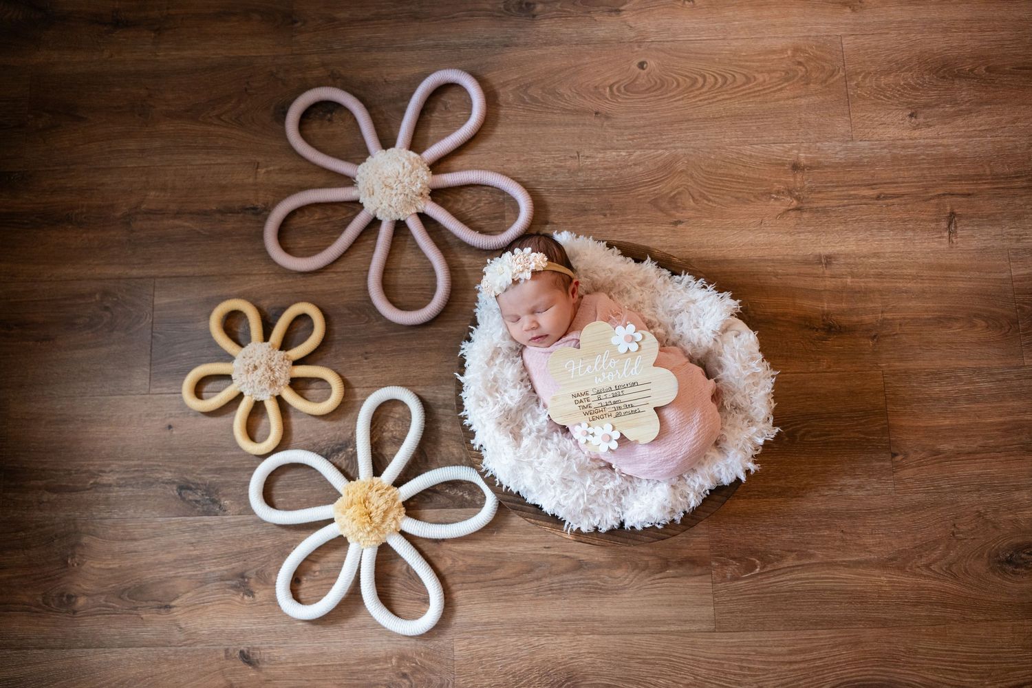 Newborn baby sleeping in fluffy basket surrounded by decorative crochet daisy flowers on wooden floor.