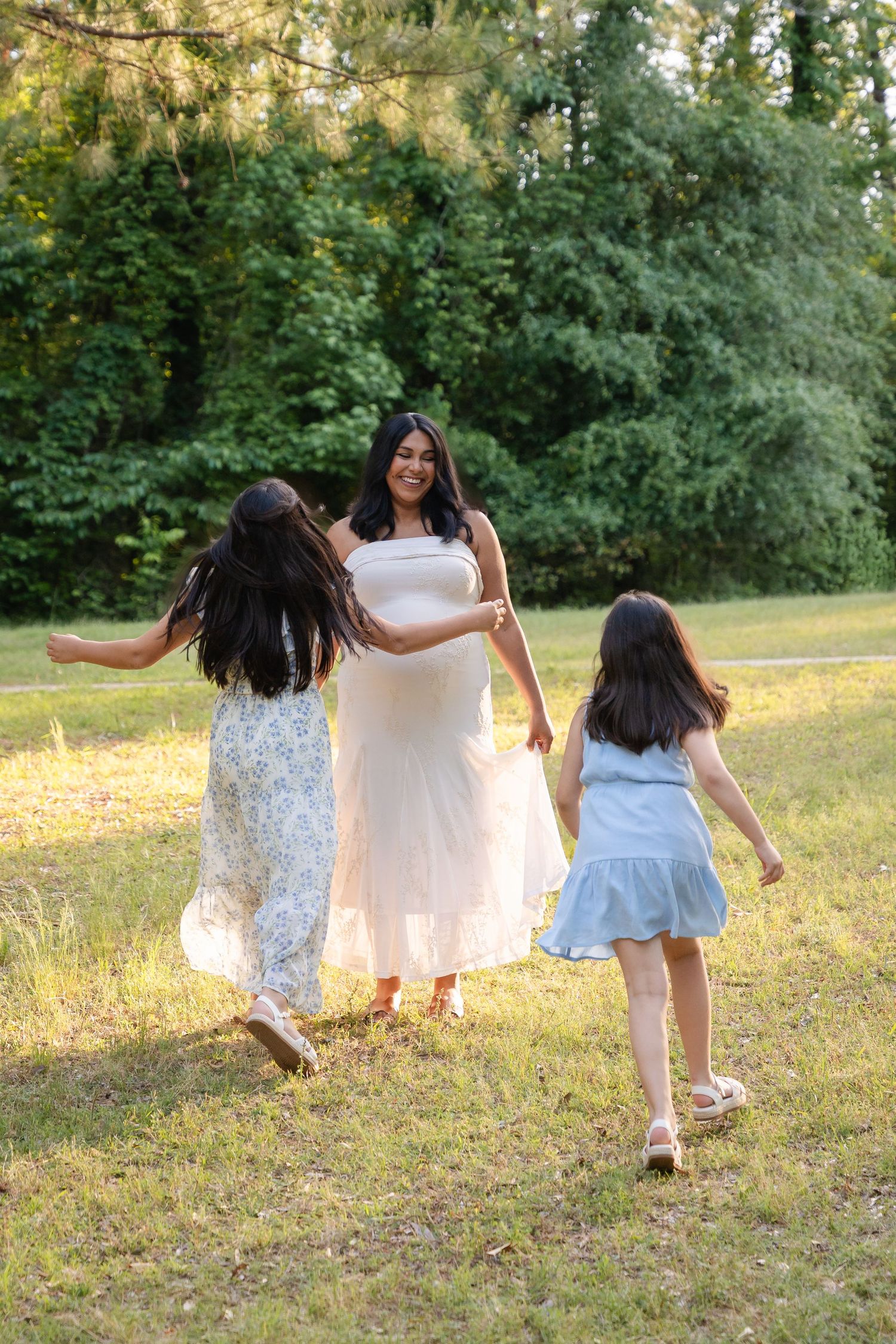 Pregnant woman in white dress laughing as two young girls run toward her in a sunny green park.