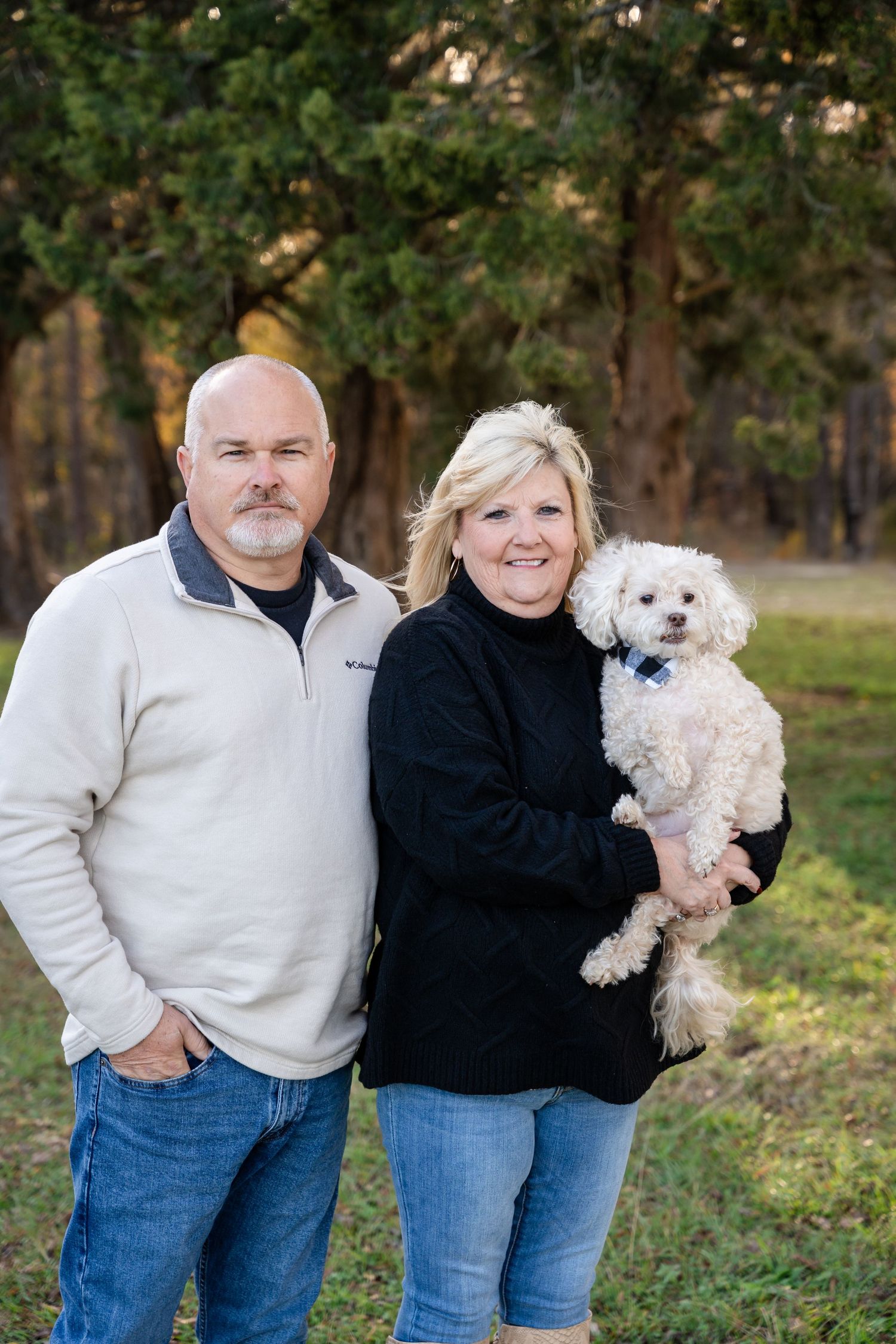 A smiling middle-aged couple stands together in a park, with the woman holding a small fluffy white dog in front of tall trees.