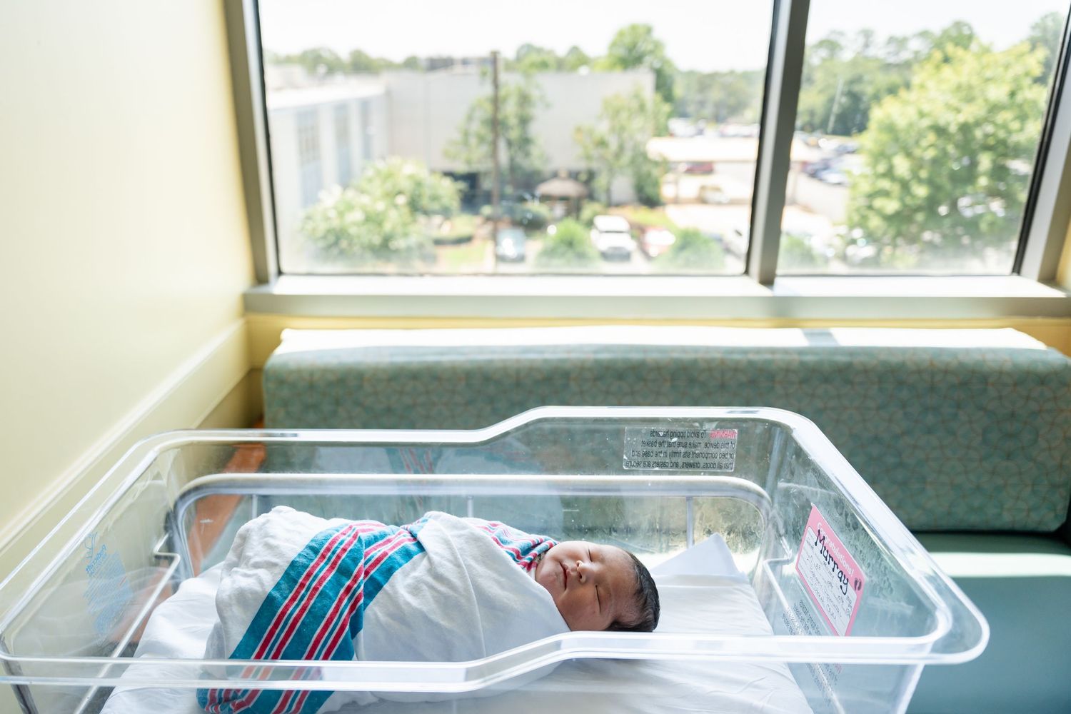 Newborn baby swaddled in a striped blanket sleeping in a clear hospital bassinet beside a sunny window overlooking trees.